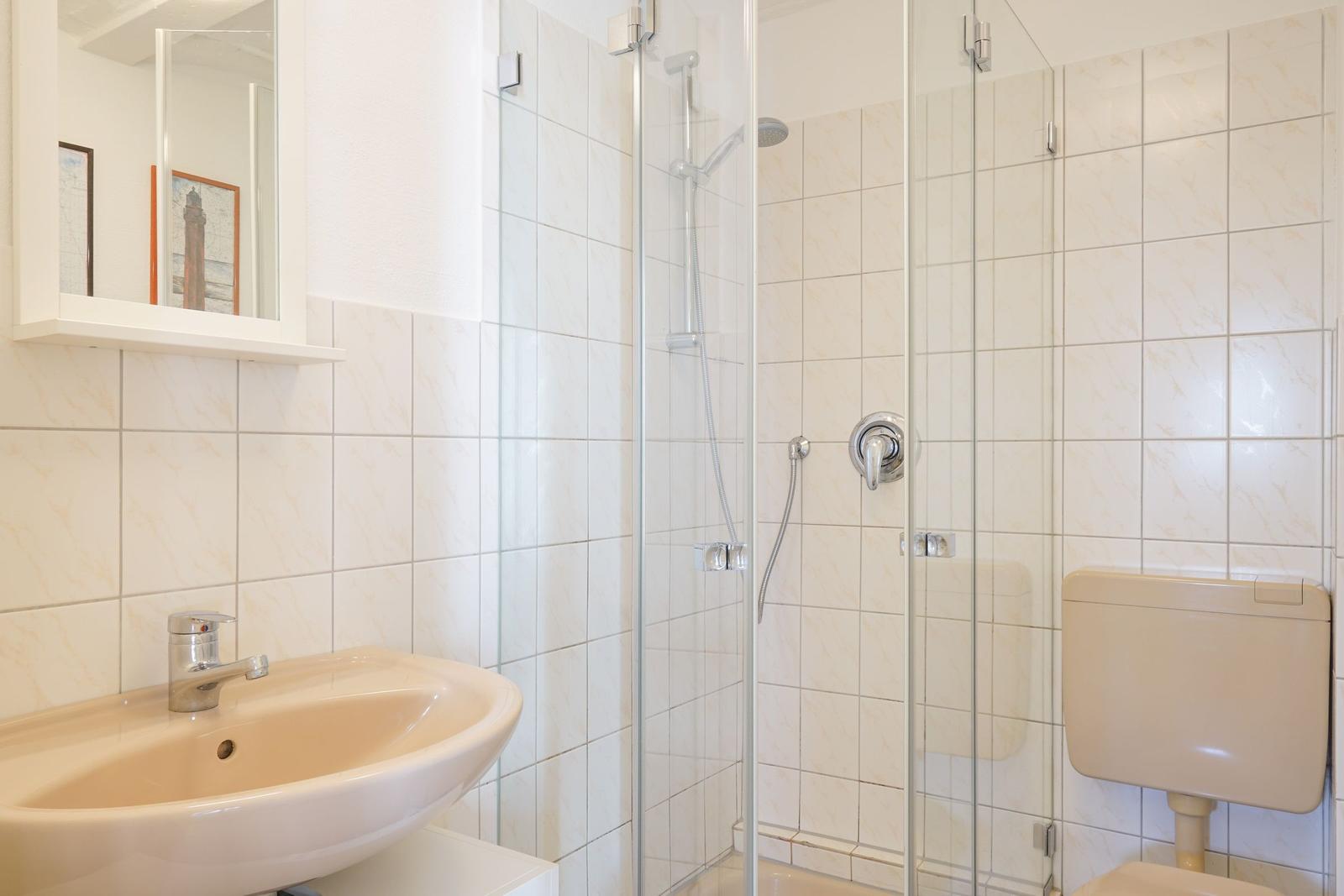 Badezimmer mit Dusche, Waschbecken und Toilette. Fliesenwand und Spiegel sichtbar.