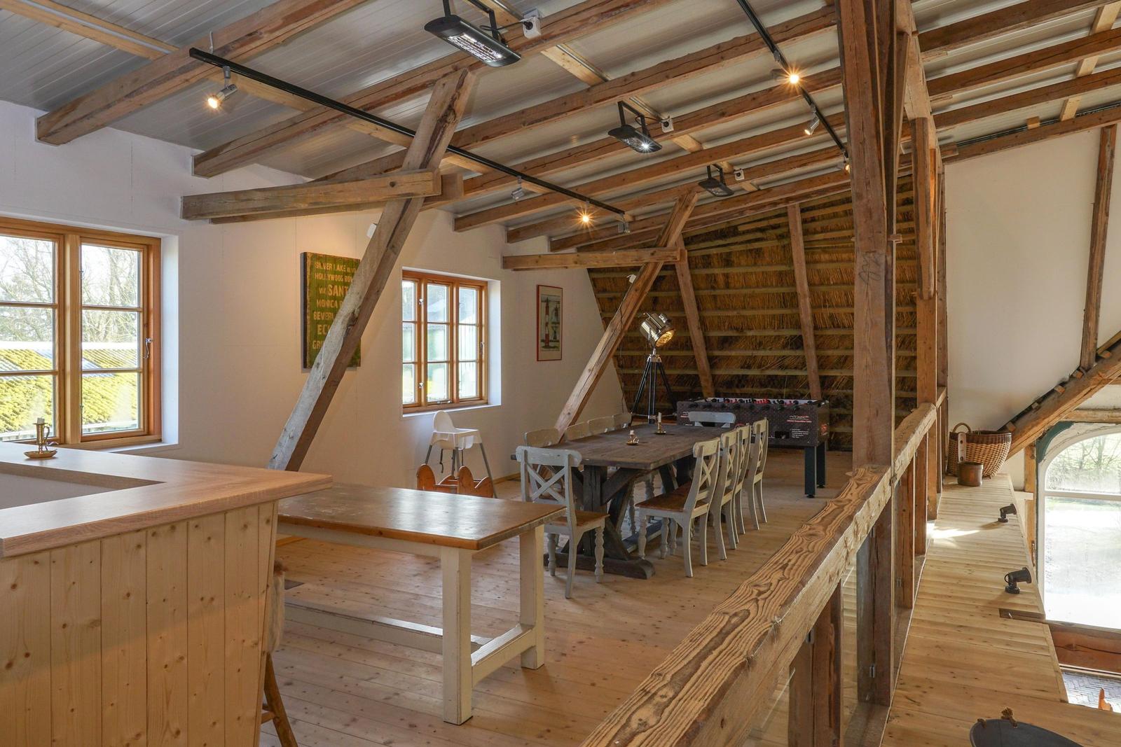 Großer Raum mit Holzdecke, Tisch, Stühle und Fenster. Dachstuhl mit Strohdecke sichtbar.