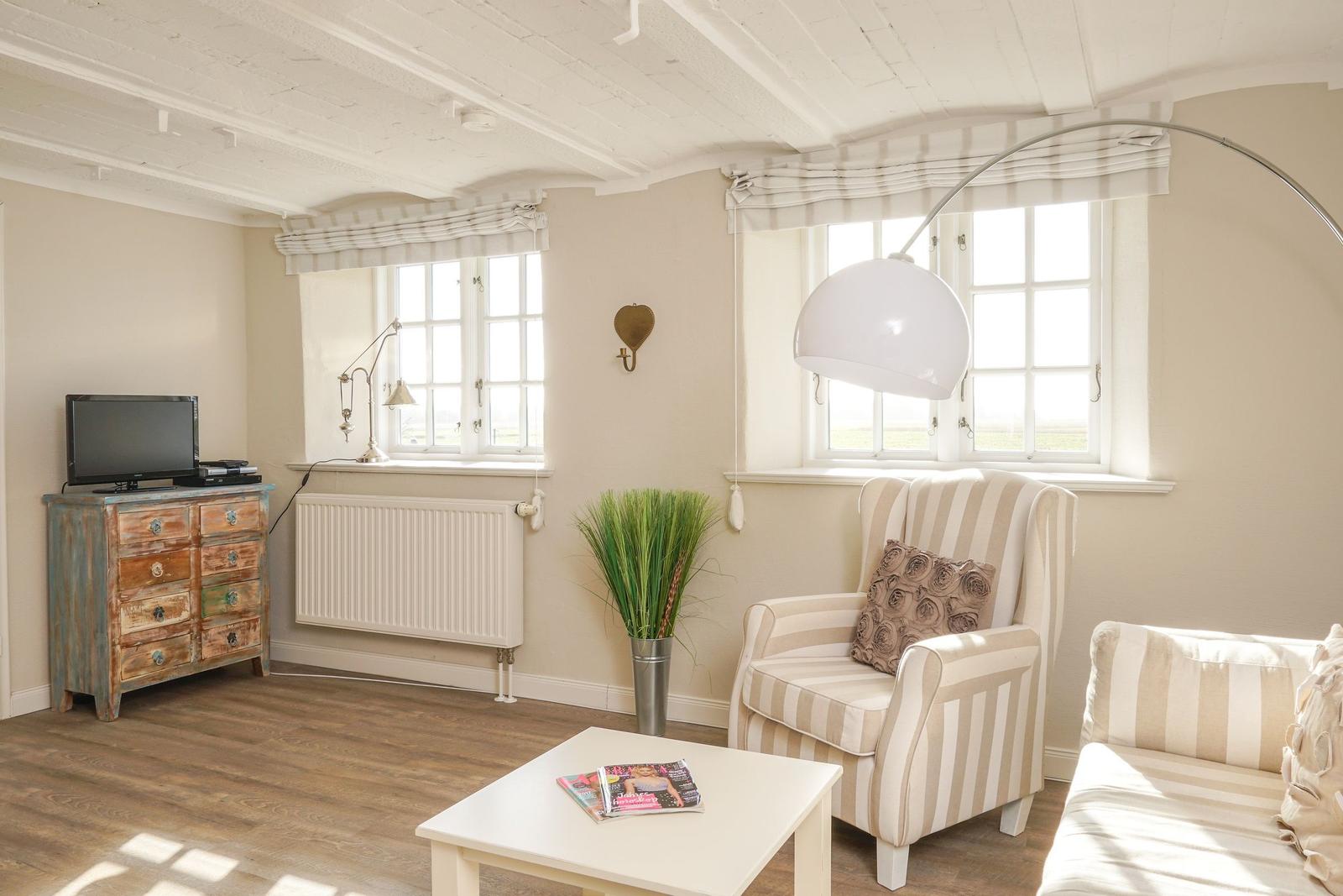 Wohnzimmer mit Sessel, Couch, TV und Fenstern. Holzboden und weiße Decke.