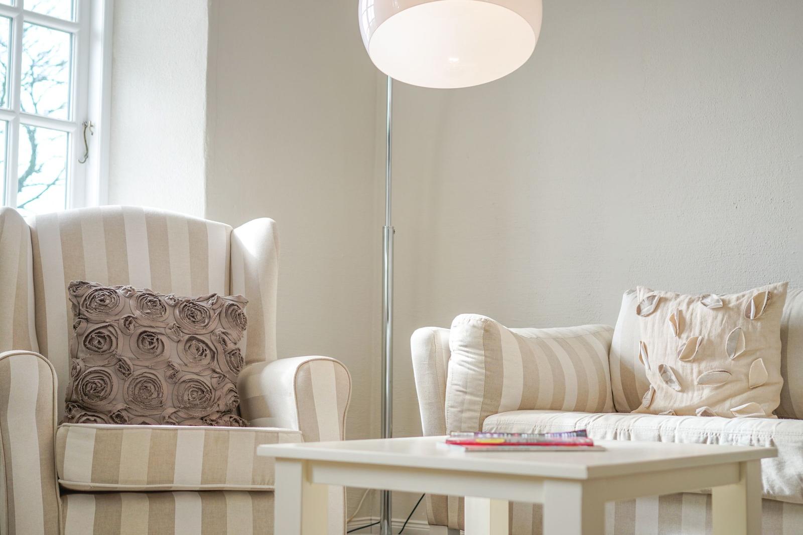 Ein Wohnzimmer mit Sessel und Sofa, Beistelltisch und Stehlampe. Fenster mit Blick nach draußen.