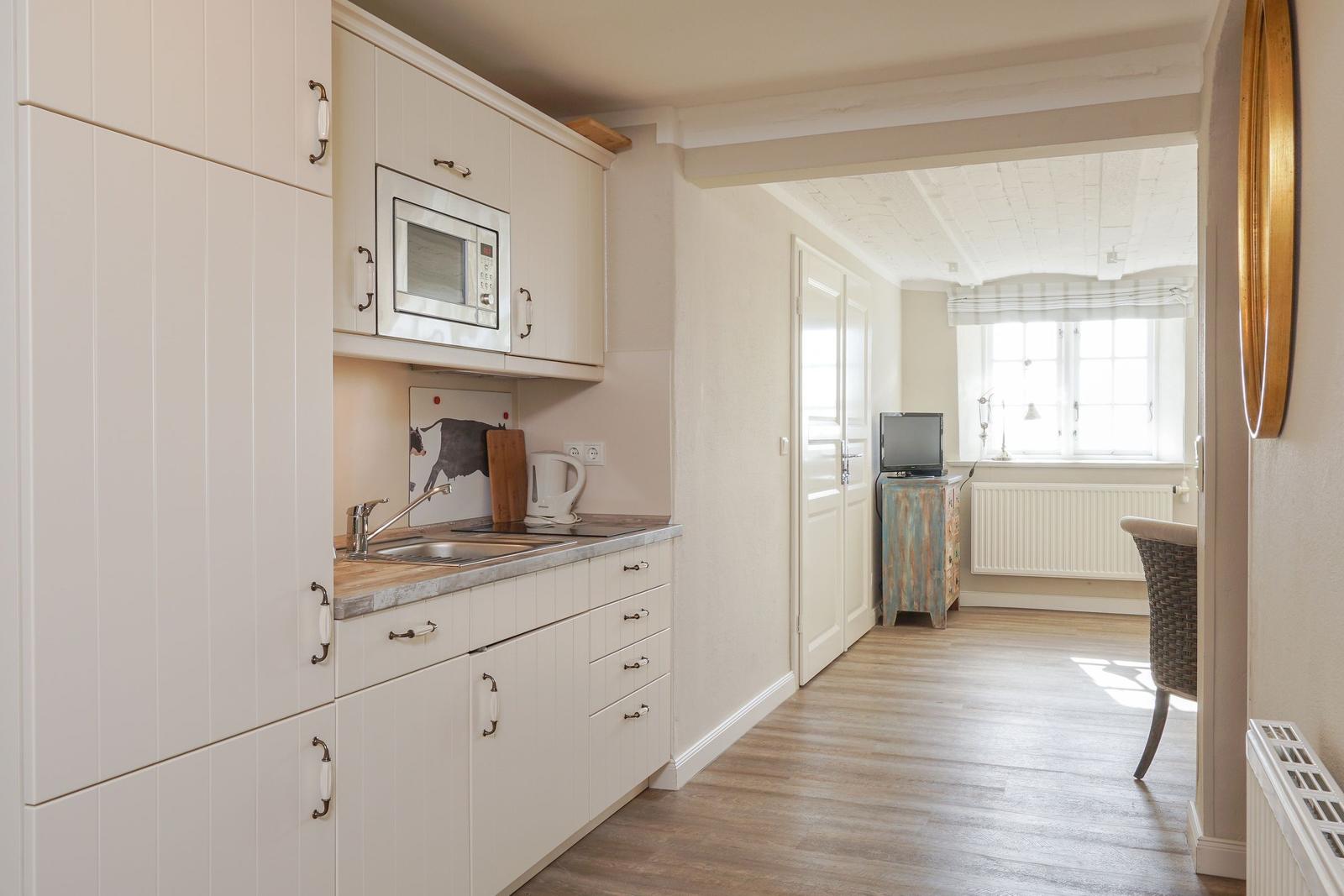 Küche mit Mikrowelle, Spüle und Arbeitsplatte. Durchgang zu Wohnzimmer mit Fenster und Stuhl.