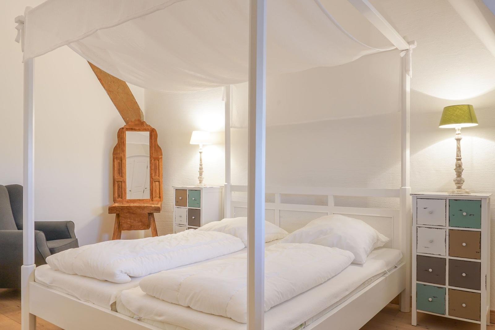Schlafzimmer mit weißem Bett, Moskitonetz, zwei Nachttischen und Holzspiegel.