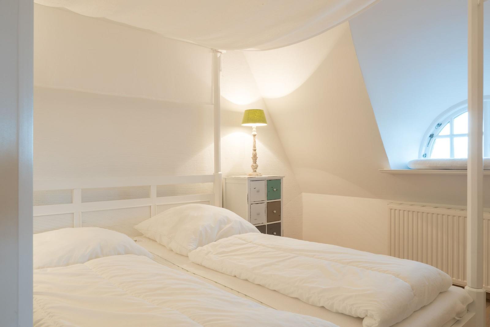 Schlafzimmer mit weißem Bett, Nachttisch und runder Fensteröffnung.