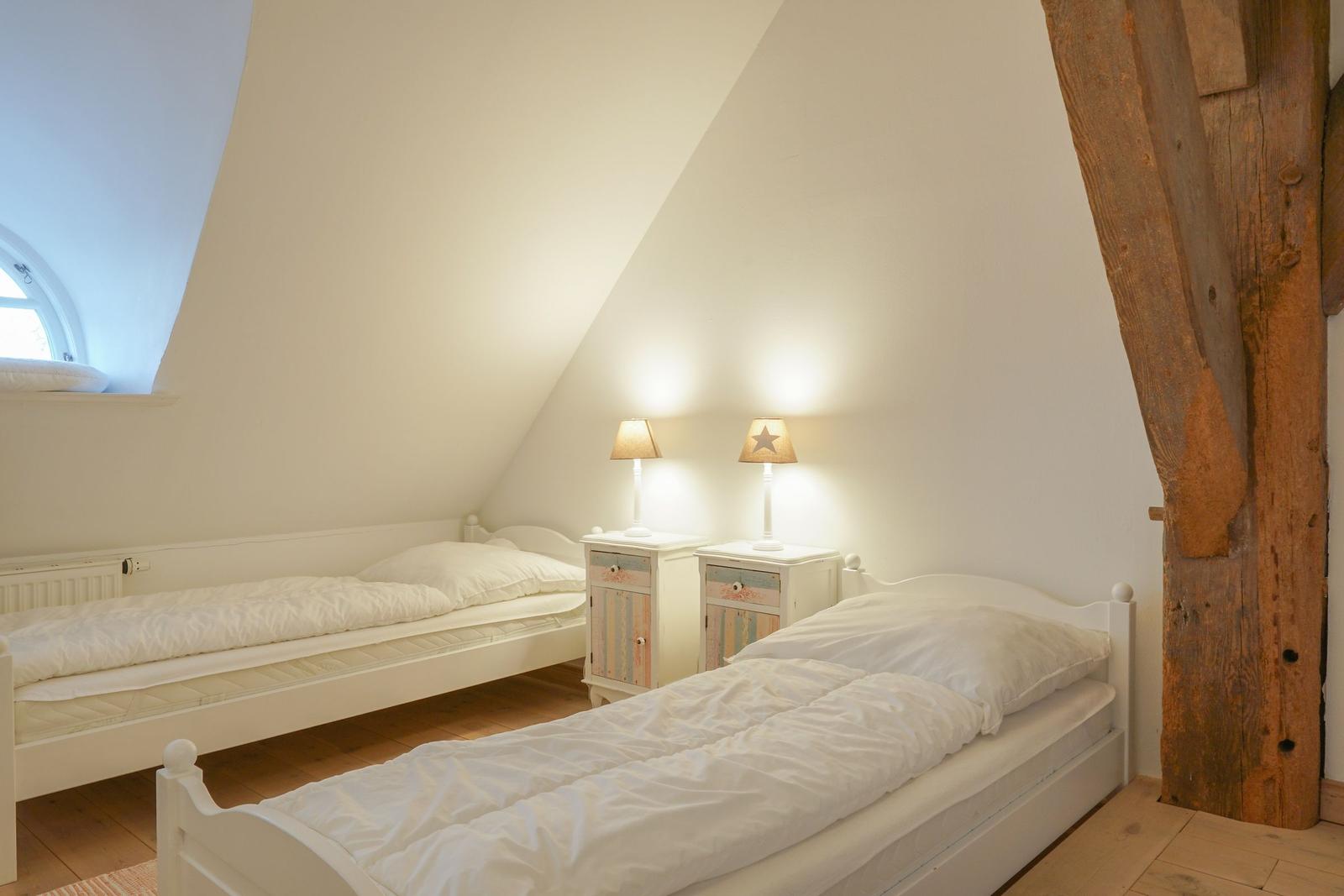 Zwei weiße Betten mit Nachttischen und Lampen unter einem Dachbodenfenster.
