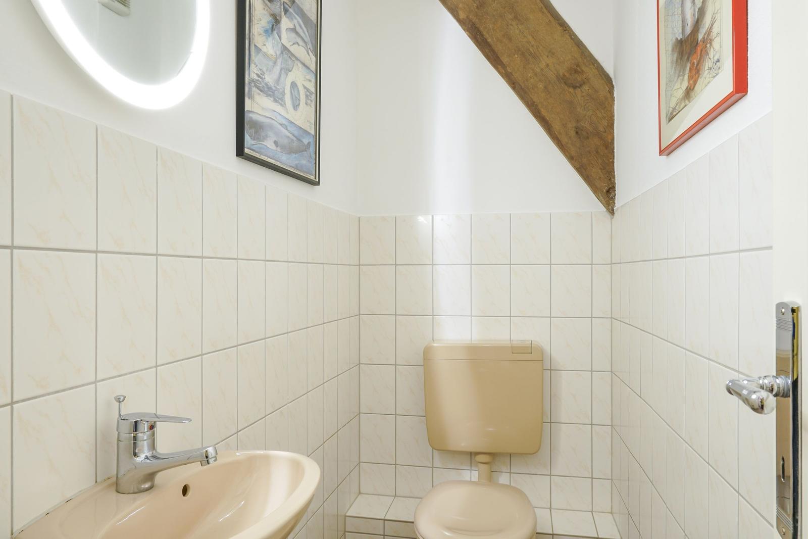 Badezimmer mit Toilette, Waschbecken und beleuchtetem Spiegel. Holzbalken an der Wand.