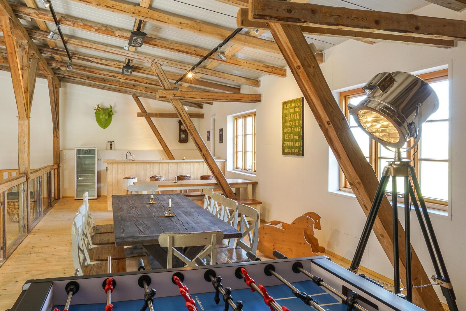 Großer Raum mit Holzdecke, Tisch, Stühle und Foosballtisch. Fenster und Lampe sichtbar.