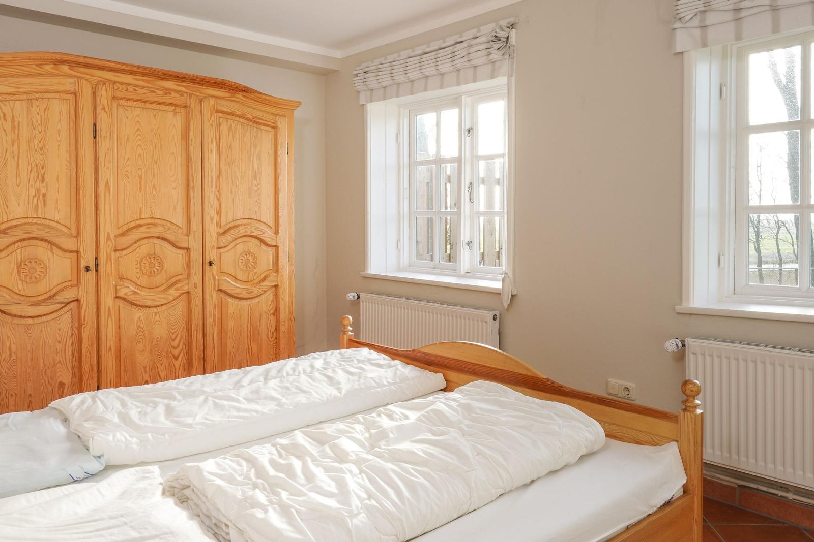 Schlafzimmer mit Holzbett, Kleiderschrank und zwei Fenstern mit Blick nach draußen.