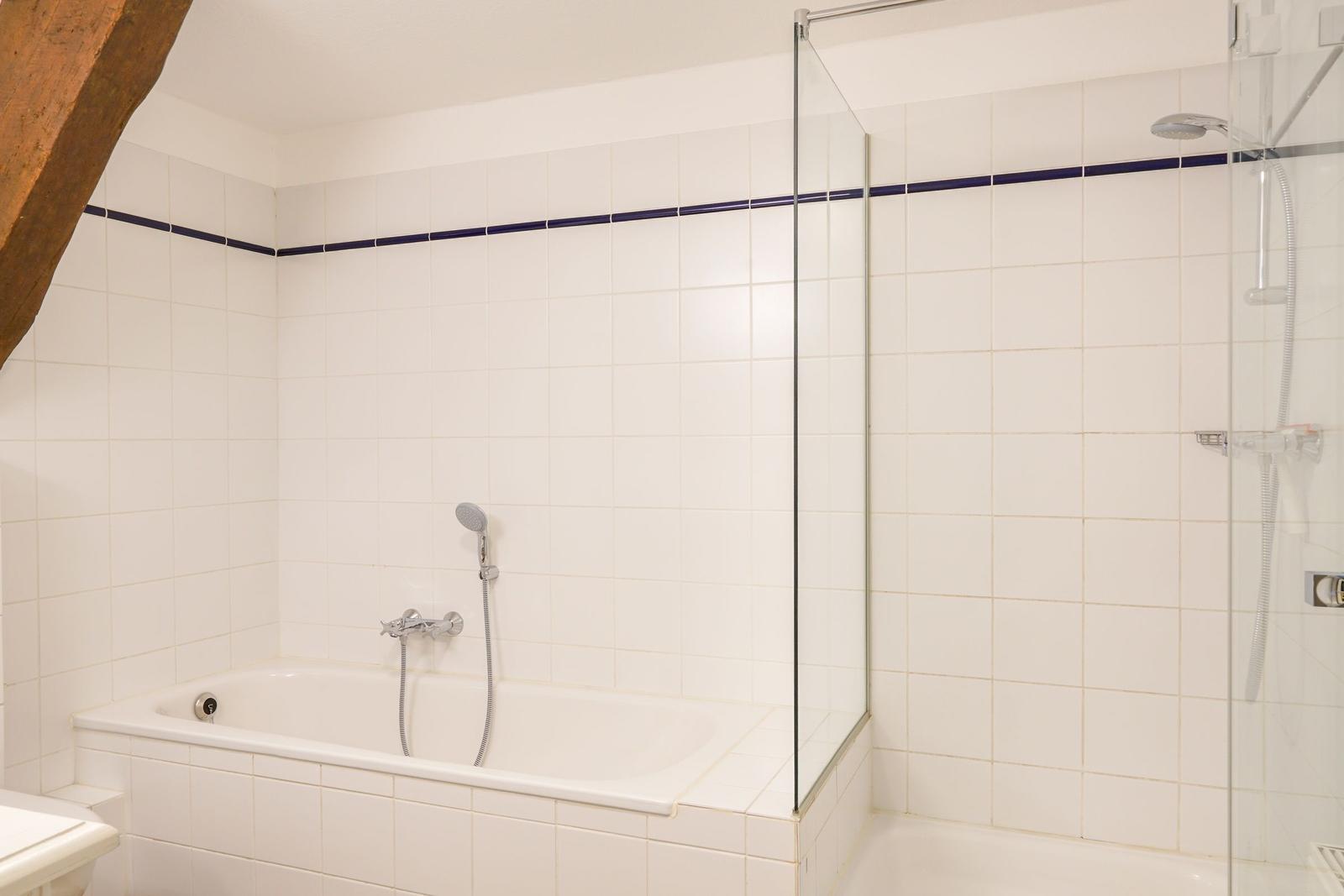 Bad mit Badewanne und Dusche, weiße Fliesen, dunkelblaue Zierleiste, Holzbalken an der Decke.