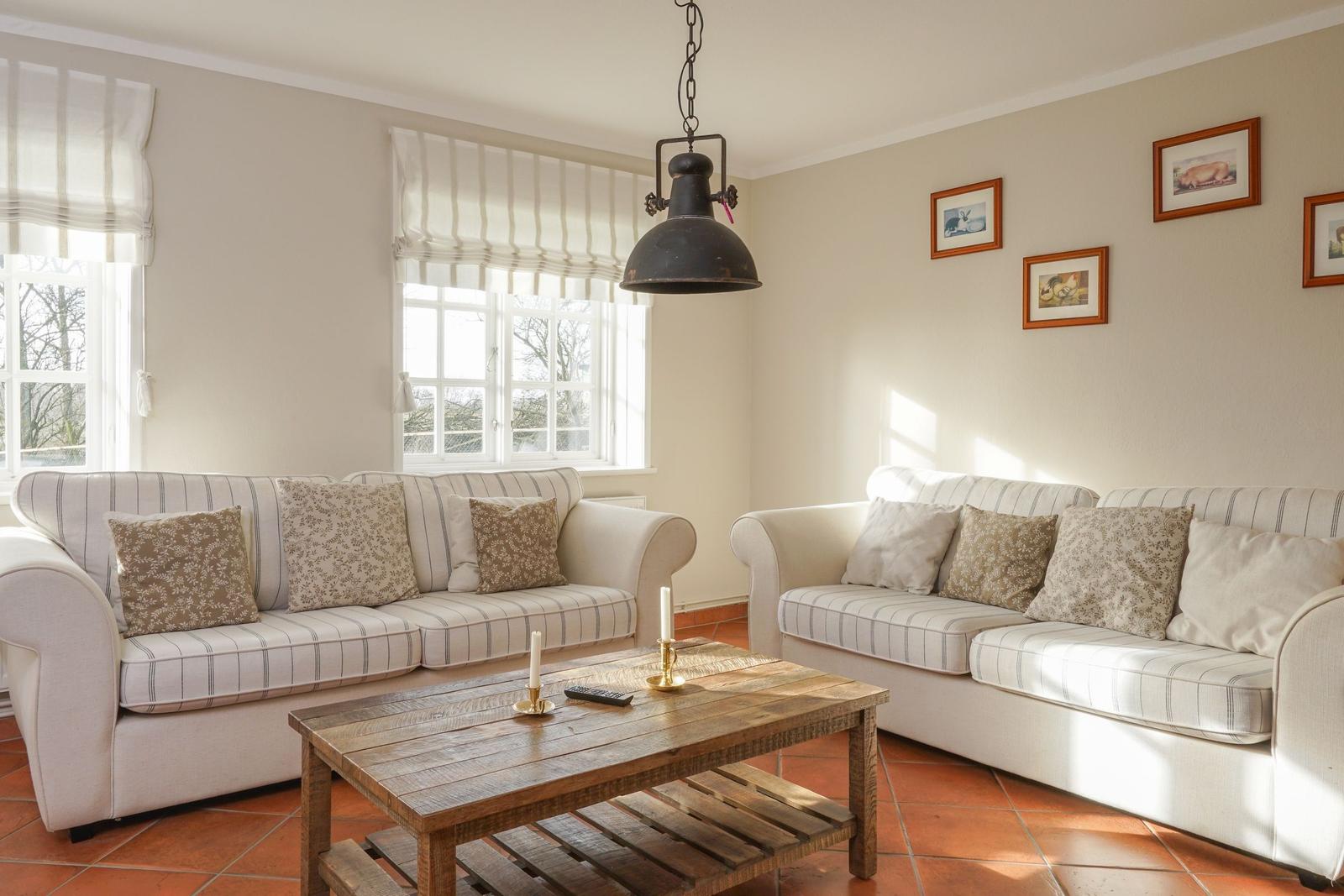 Zwei Sofas mit Streifenbezug, Holztisch und Wandbilder in einem Wohnzimmer mit Fenstern und Terrakottaboden.