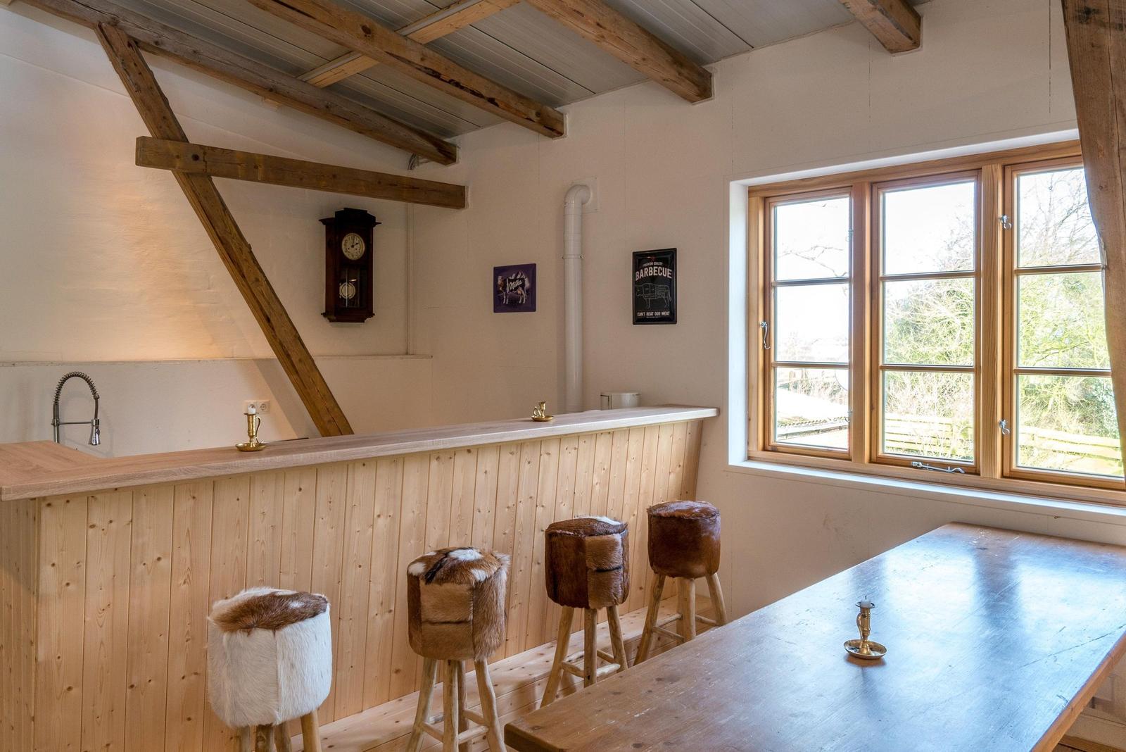 Raum mit Holzbar, Stühlen und Fenster. Deckenbalken und Wanduhr sichtbar.