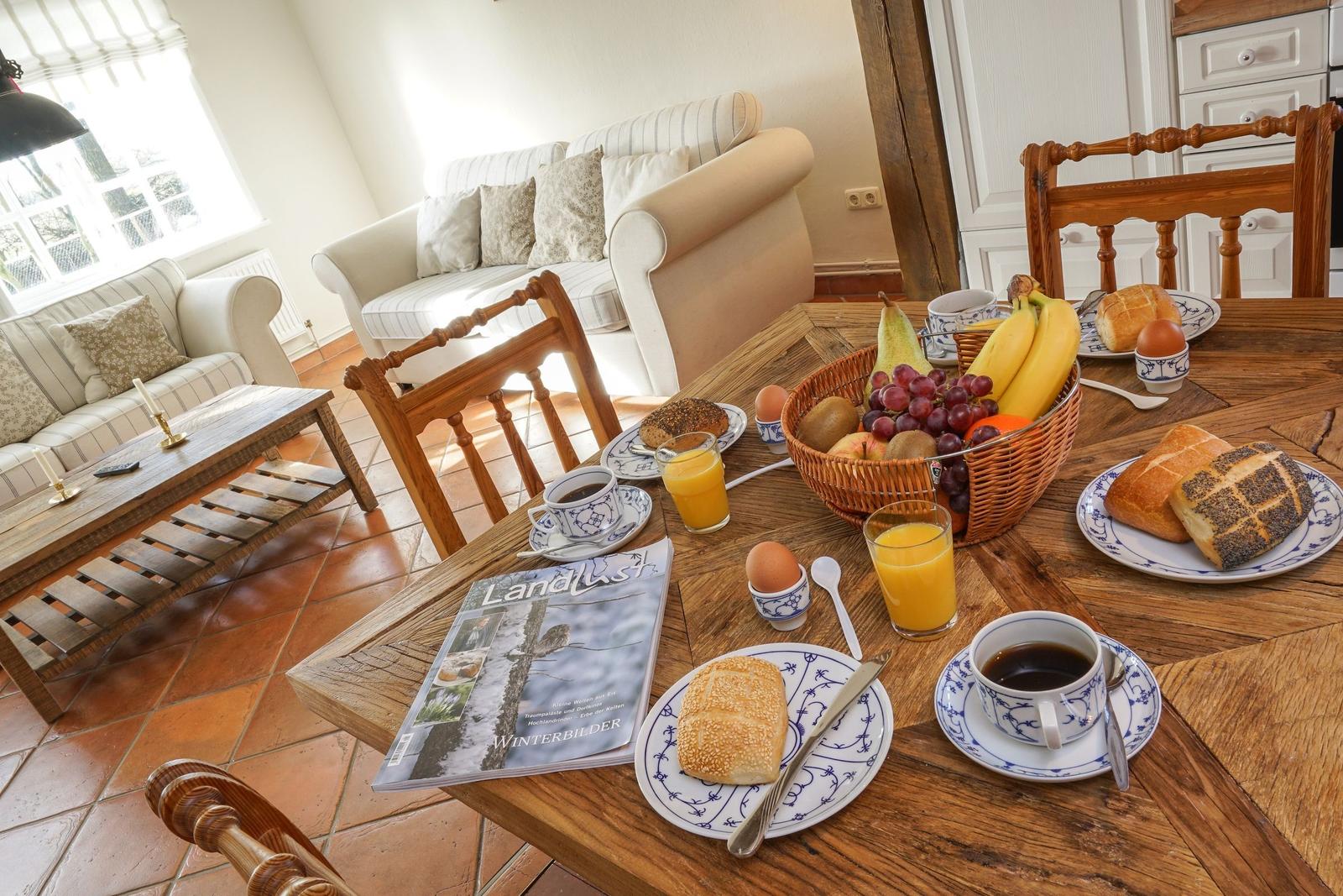 Frühstückstablett mit Obst, Brot und Getränken auf Holztisch. Im Hintergrund Sofa und Fenster.