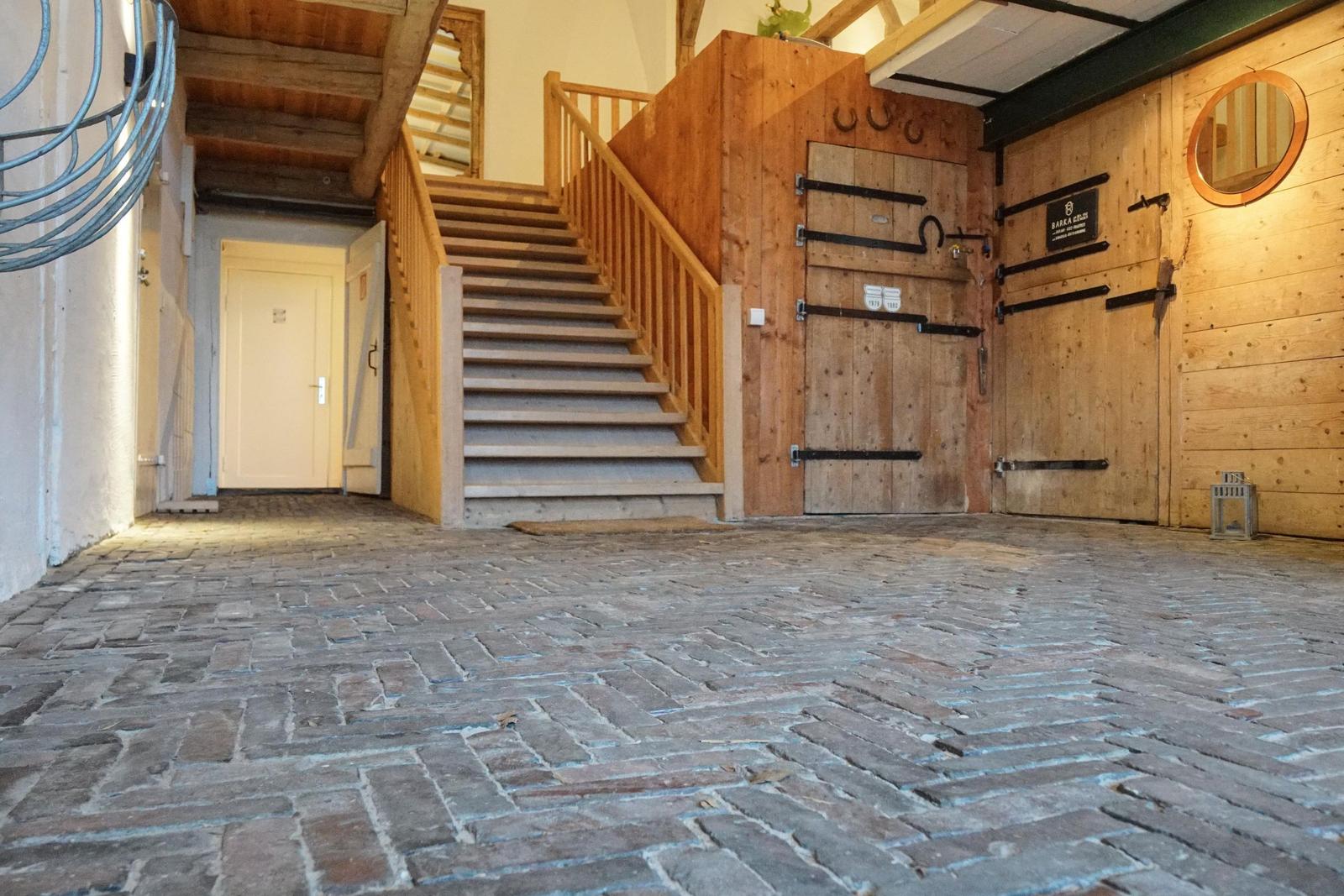 Helle Eingangshalle mit Holztreppe und Steinboden. Holzwände und Türen mit Metallbeschlägen.