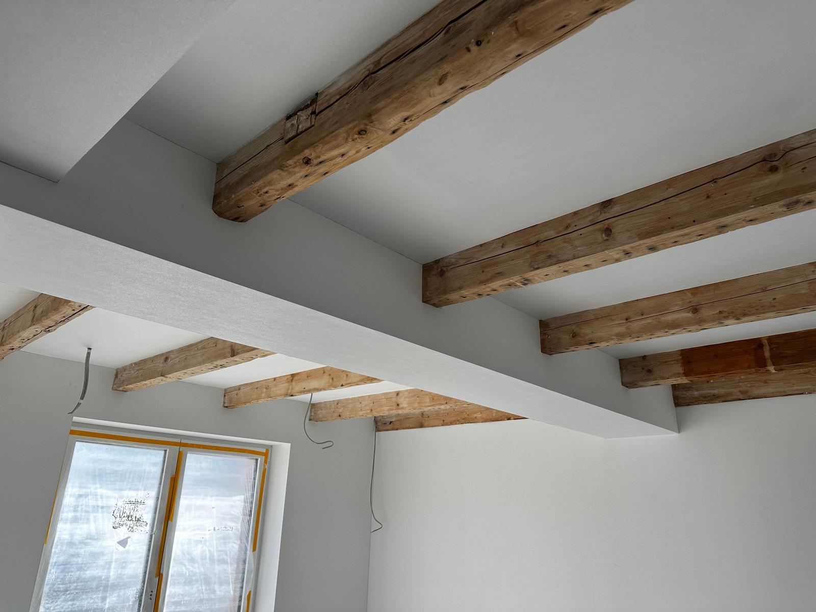 Raum mit sichtbaren Holzbalken an der Decke und grauen Wänden. Fenster mit gelber Schutzfolie.