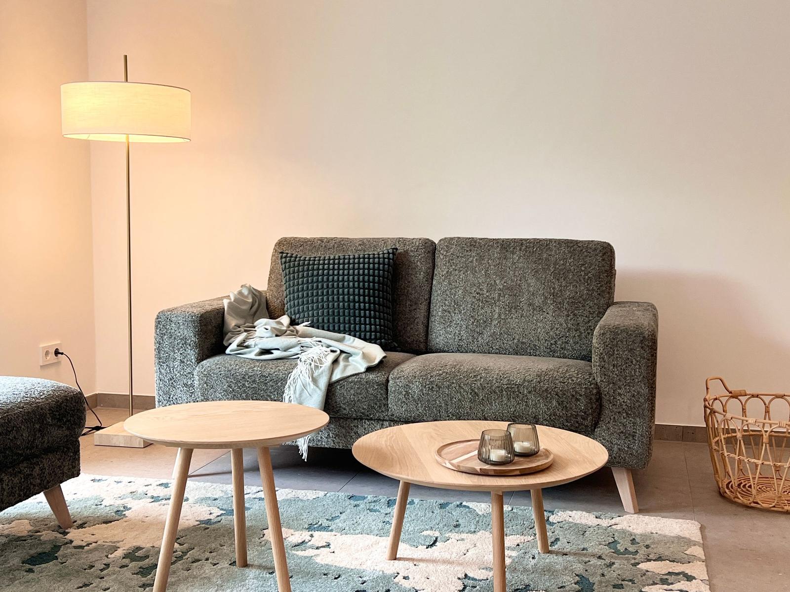 Ein grauer Sofa mit Kissen und Decke, zwei runde Holztische und ein Stehlampe.