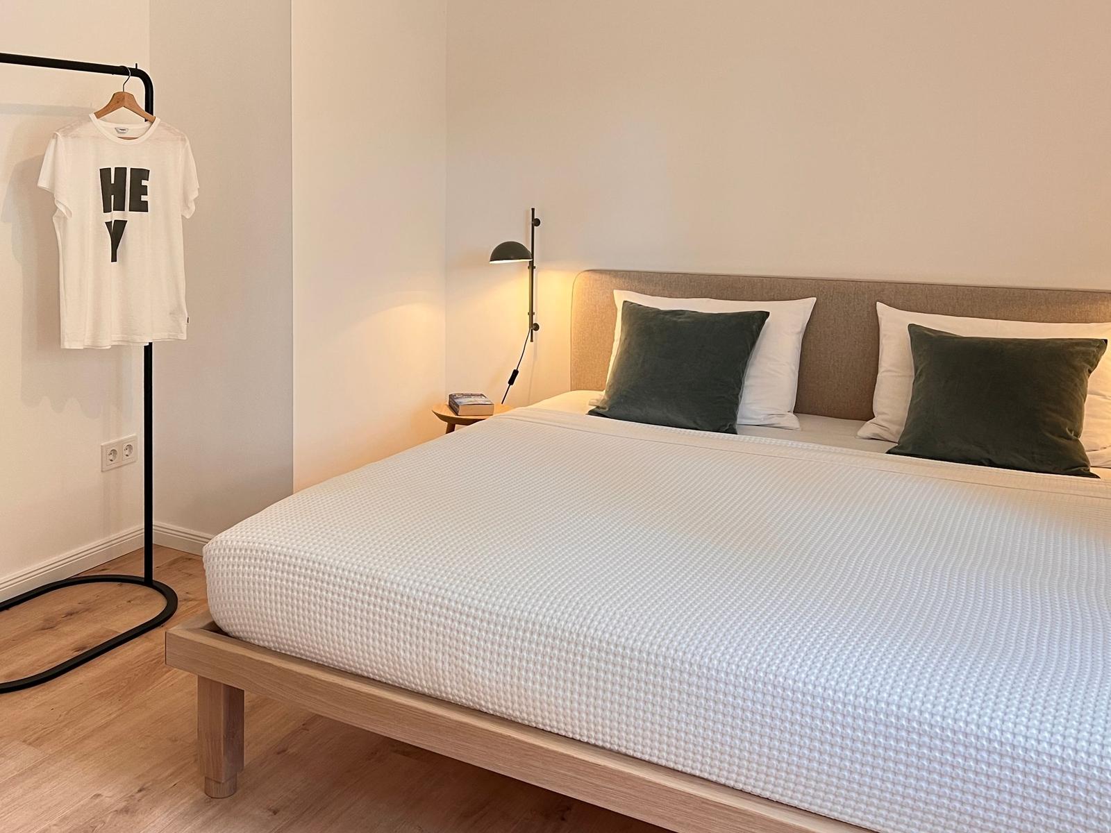 Ein Bett mit weißer Bettwäsche und grünen Kissen. Eine Lampe und ein Kleiderständer sind sichtbar.