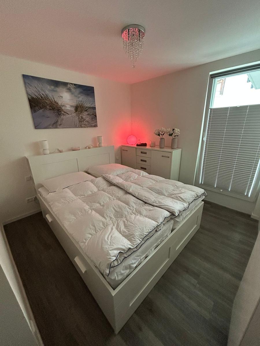 Schlafzimmer mit weißem Bett, Wandbild und Fenster mit Jalousie.