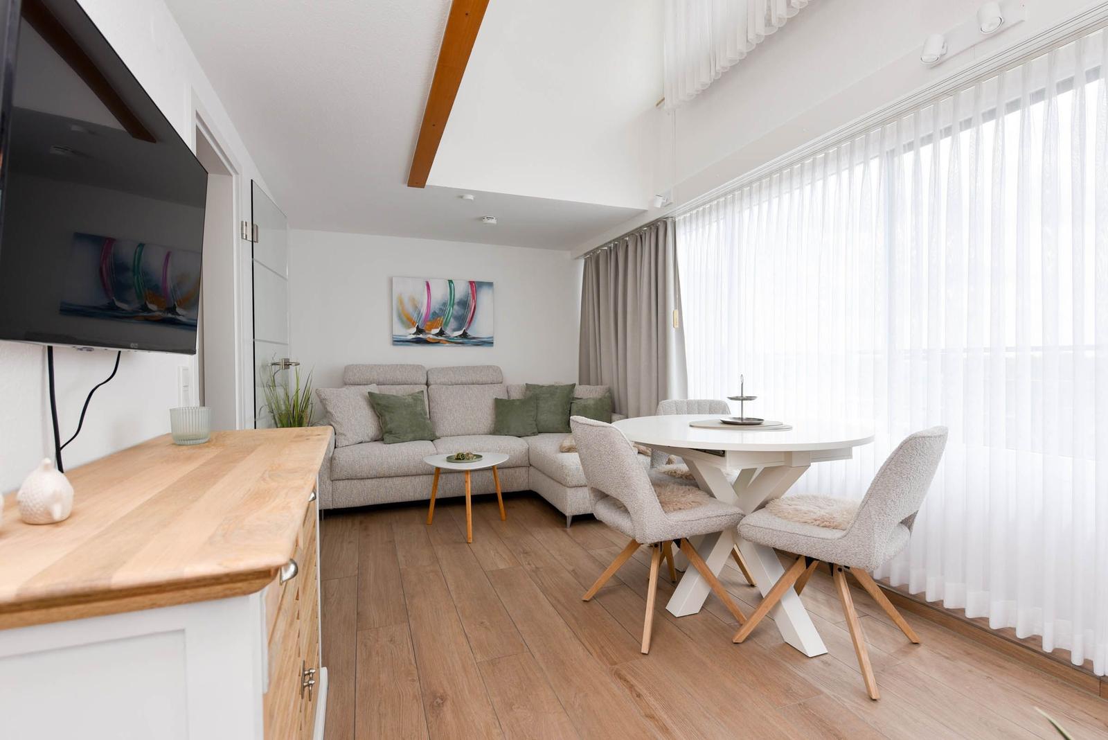 Wohn- und Essbereich mit Sofa, runden Tisch und TV. Holzfußboden und große Fenster.