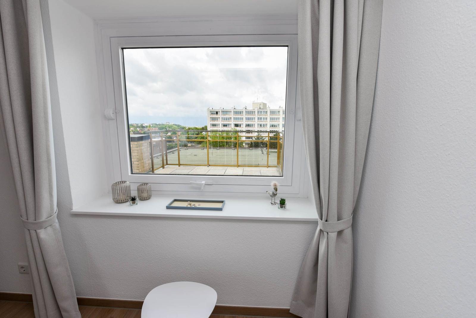 Ein Fenster mit Blick auf eine Terrasse und ein Gebäude. Vor dem Fenster stehen dekorative Gegenstände.