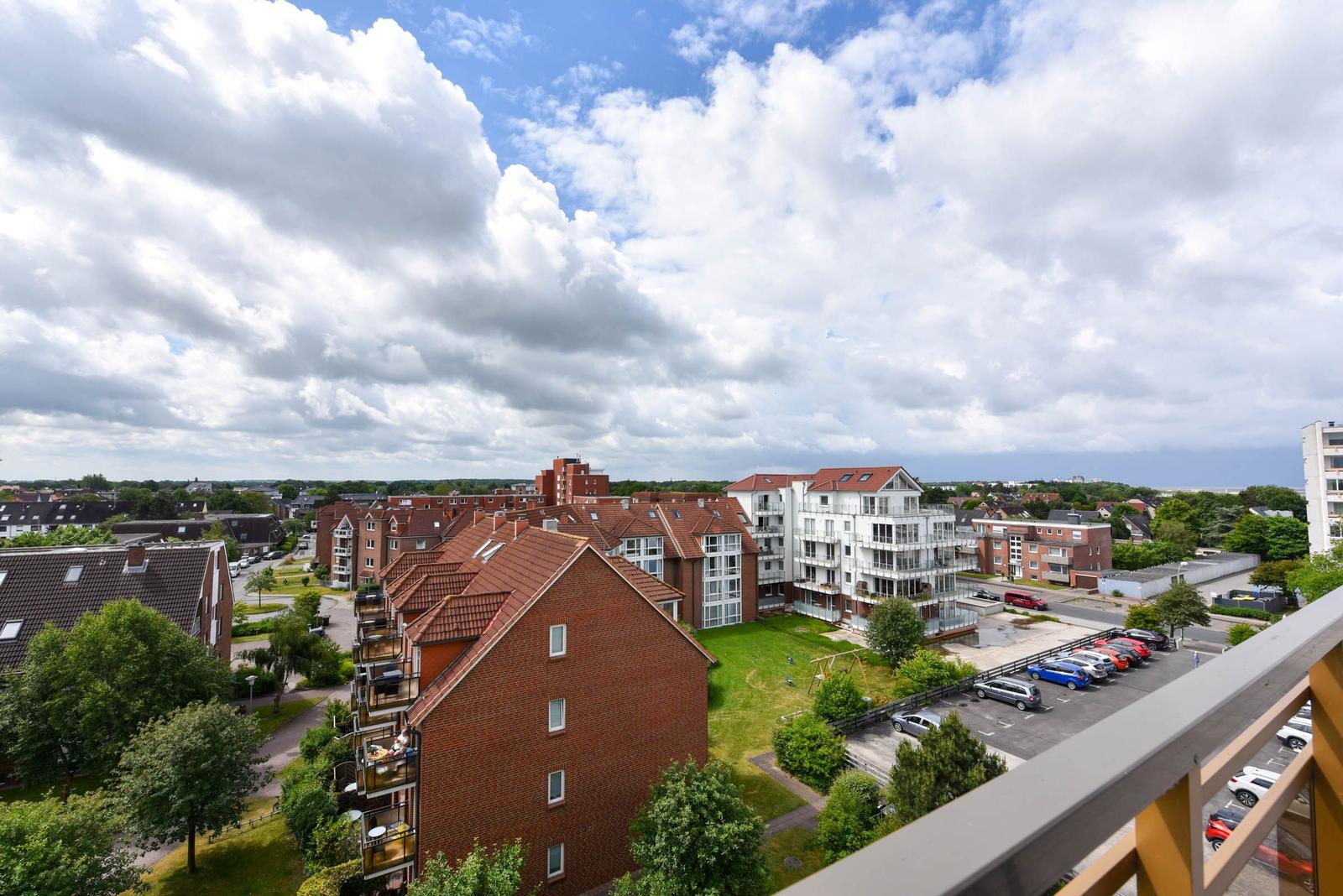 Blick von Balkon auf Wohngebiet mit Häusern, Grünflächen und Parkplatz unter blauem Himmel mit Wolken.