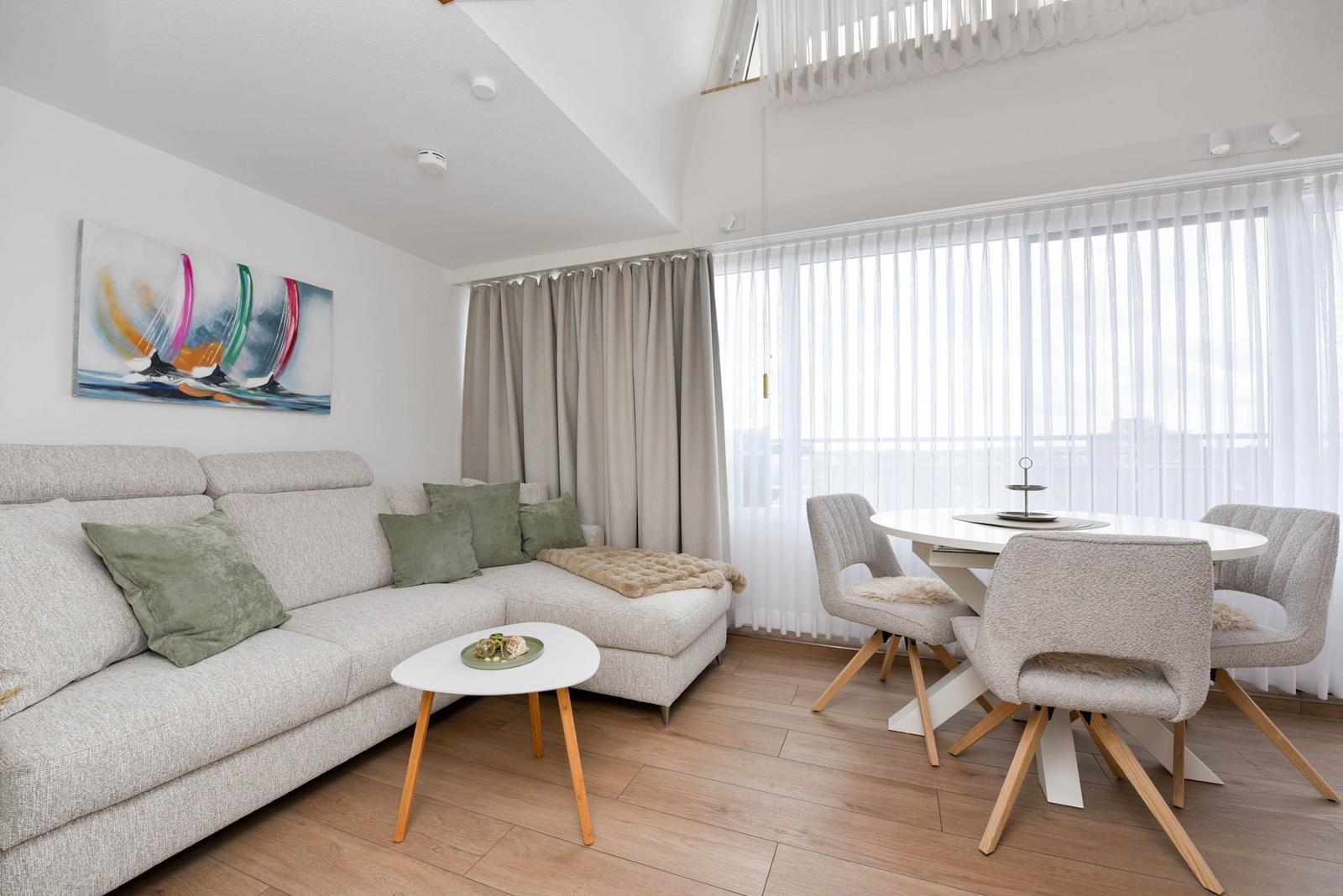 Wohn- und Essbereich mit Sofa, Tisch und Stühlen. Große Fenster mit weißen Vorhängen.