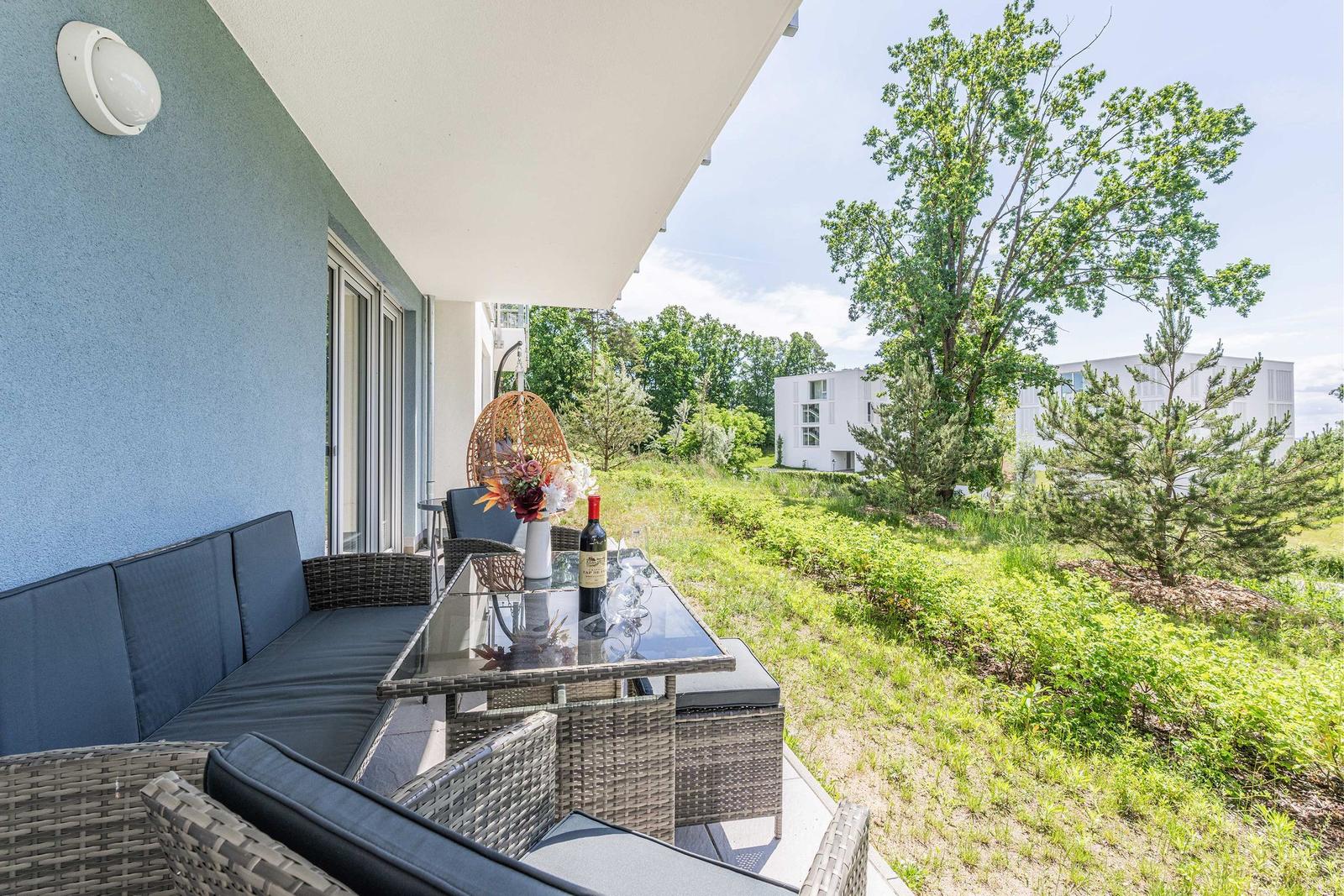 Terrasse mit Sitzgruppe, Tisch und Weinflasche. Grünfläche und Bäume im Hintergrund.
