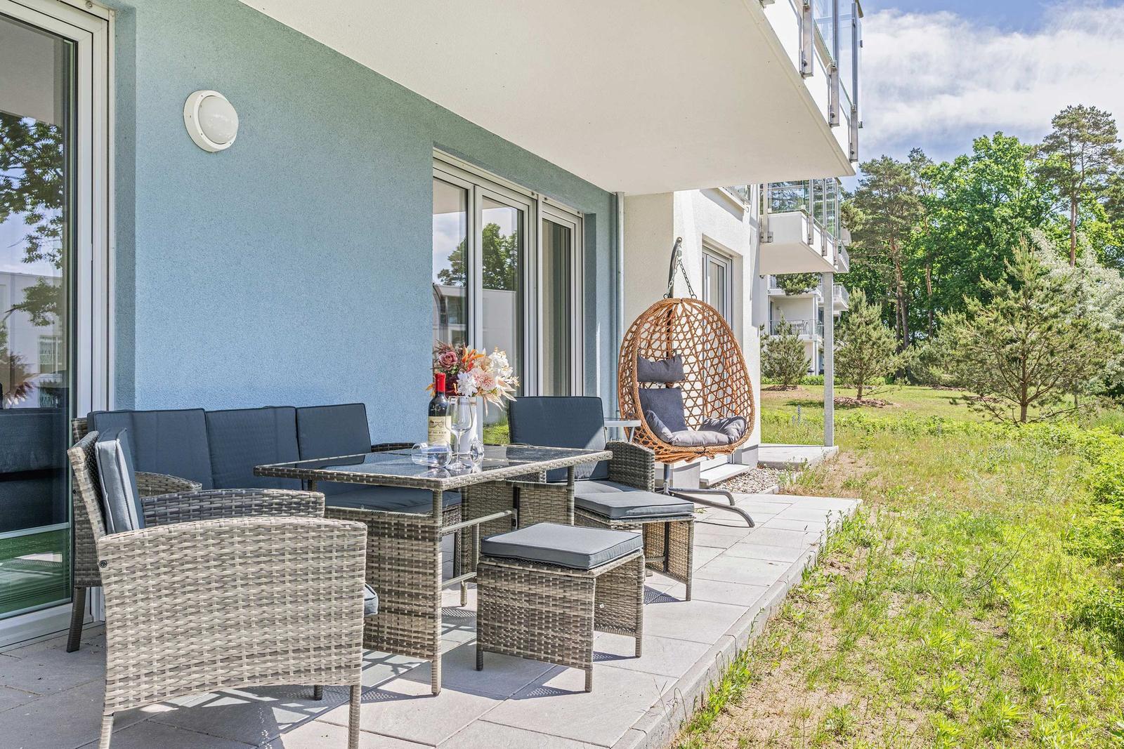 Terrasse mit Sitzgruppe, Esstisch und Hängesessel. Grünfläche und Bäume im Hintergrund.
