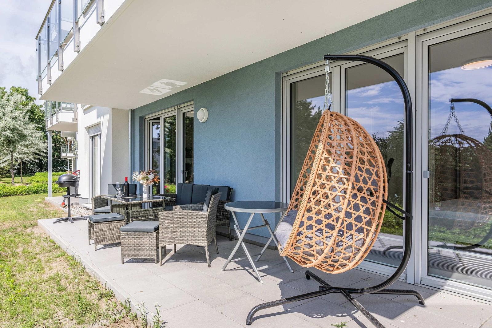 Terrasse mit Hängesessel, Sitzgruppe und Grill. Glasfront und blauer Putz sichtbar.