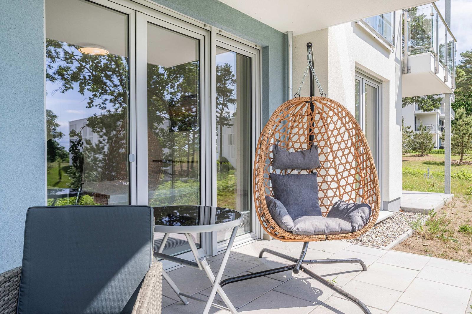 Terrasse mit Hängesessel, Tisch und Stuhl. Glasfront und Garten im Hintergrund.