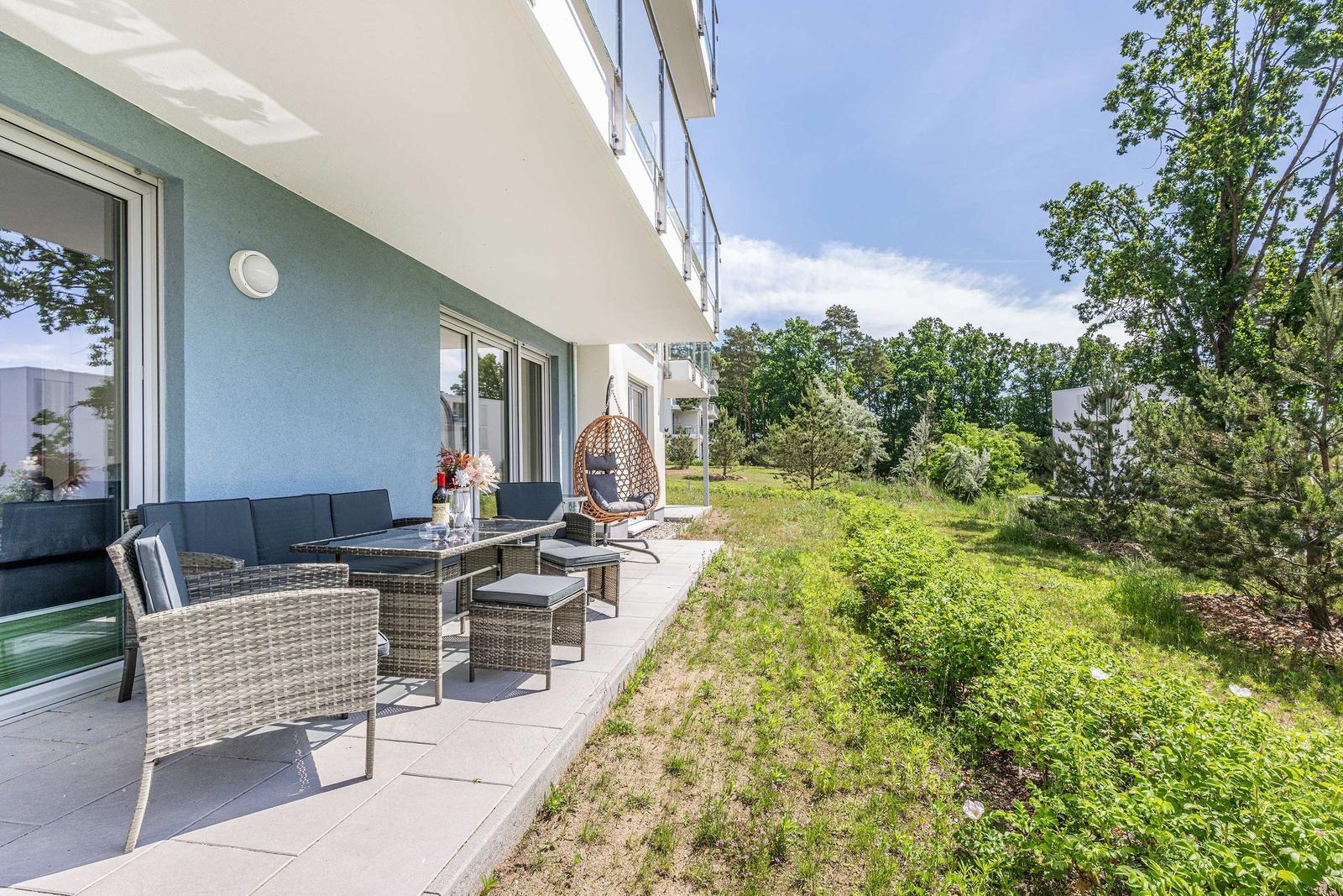 Terrasse mit Sitzgruppe, Tisch und Hängesessel. Grünflächen und Bäume im Hintergrund.