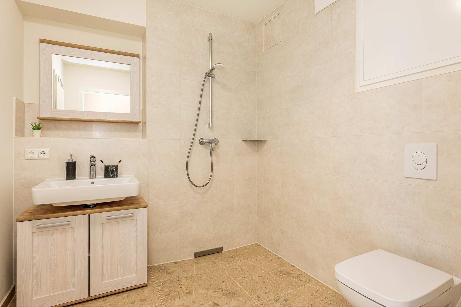 Badezimmer mit Dusche, Waschbecken und Toilette. Wand- und Bodenfliesen in hellen Tönen.