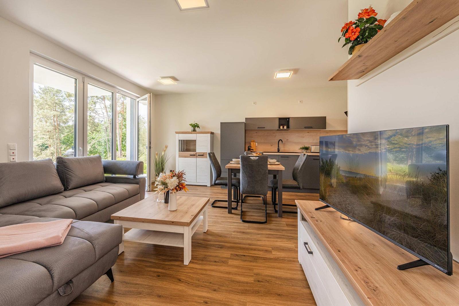 Wohn- und Essbereich mit Sofa, TV und Küchenzeile. Holzboden und große Fenster.