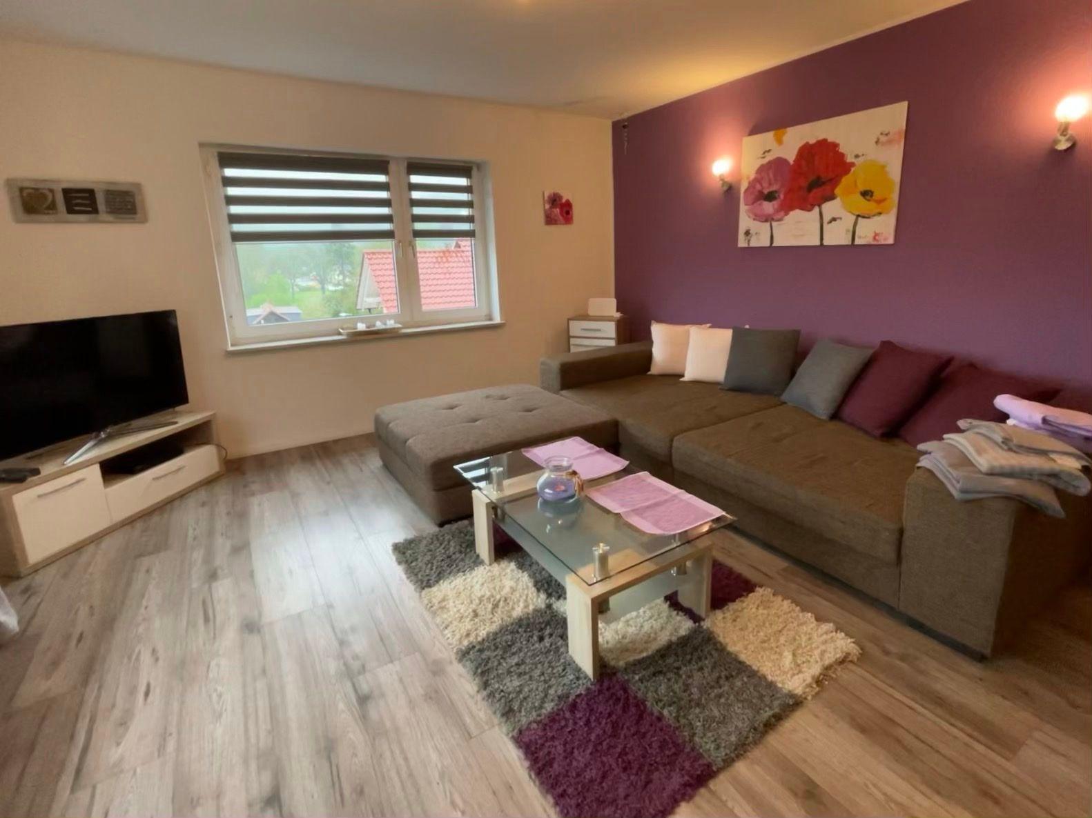 Wohnzimmer mit Sofa, Glas-Tisch und Fernseher. Wand in Lila mit Blumenbild.