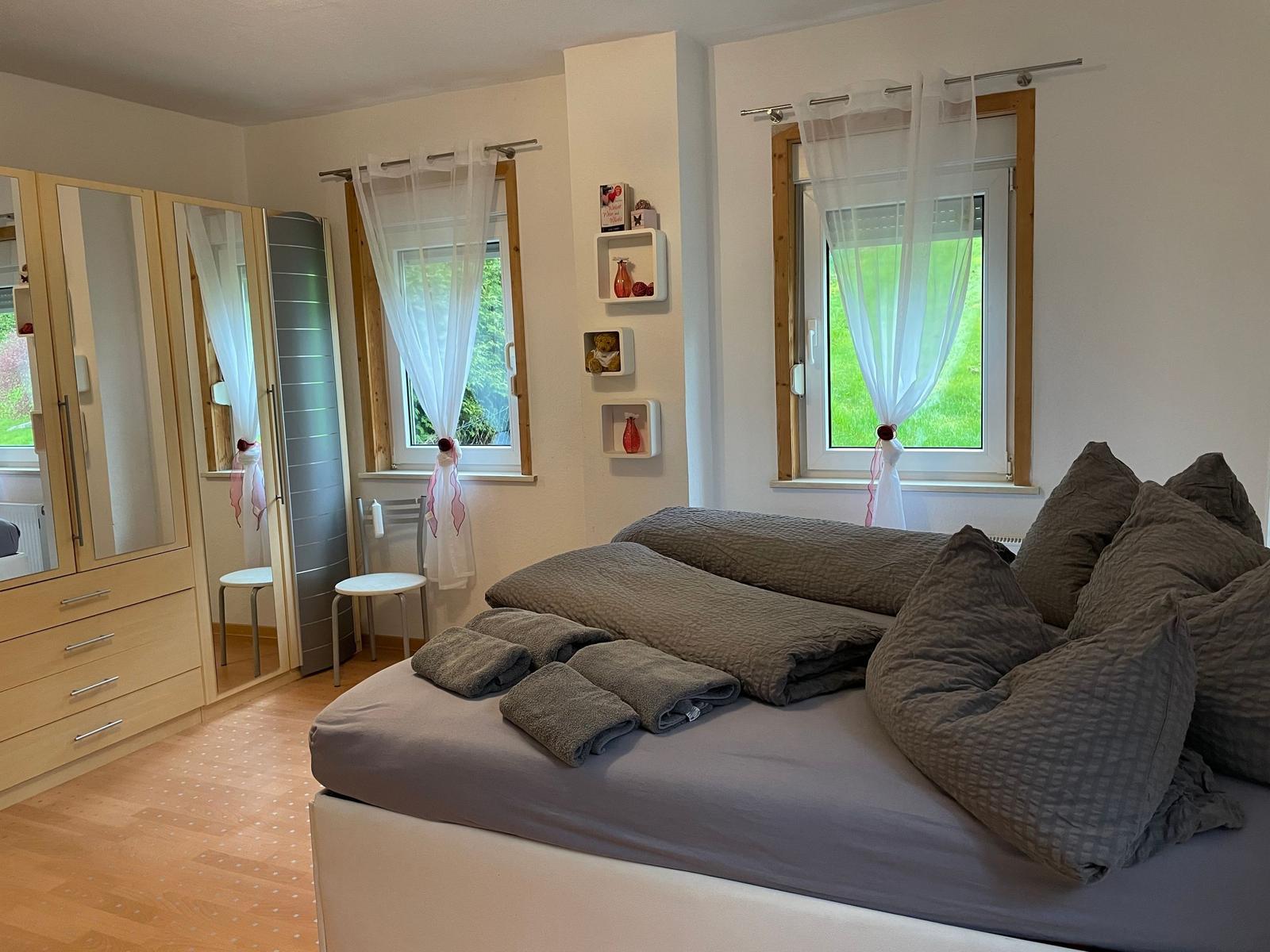 Schlafzimmer mit Doppelbett, Kleiderschrank und zwei Fenstern mit weißen Vorhängen.