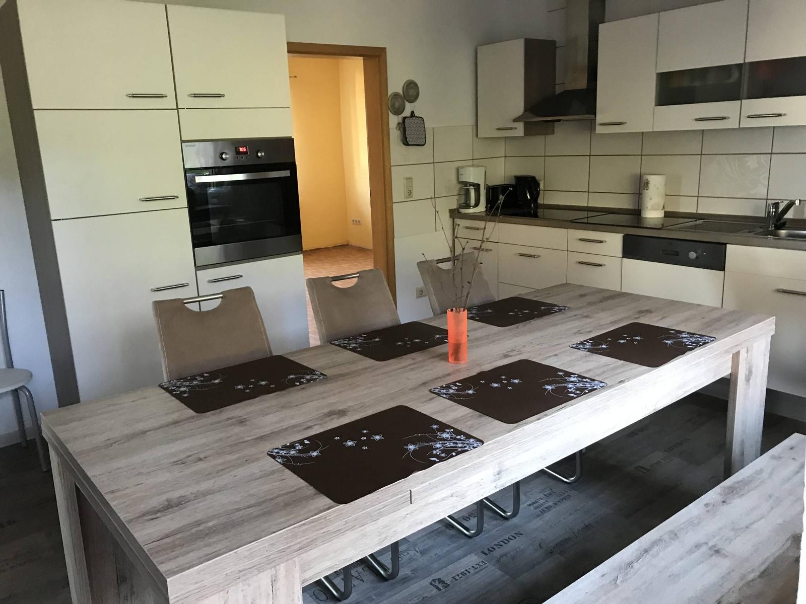 Essbereich mit Holztisch und Stühlen. Küche mit weißen Schränken und Einbauküche.