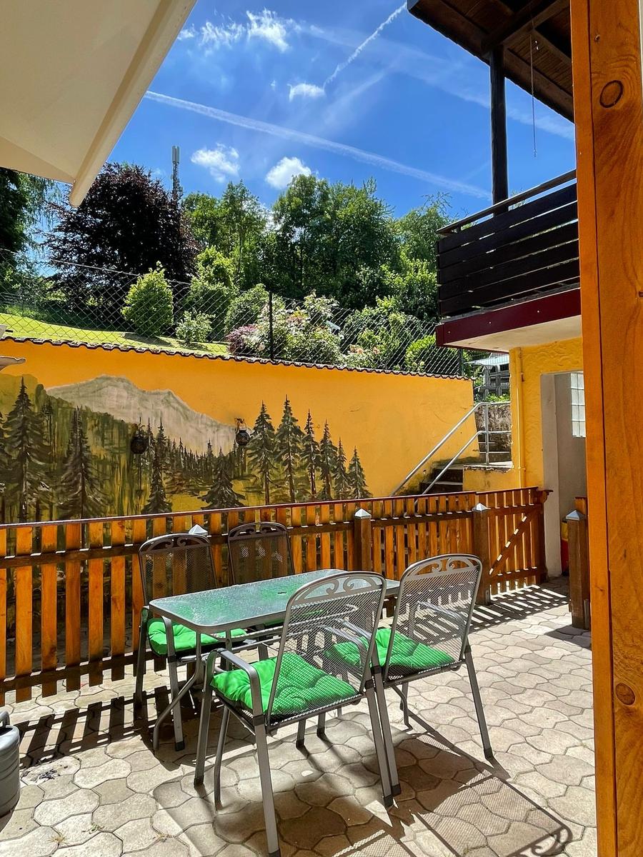Terrasse mit Tisch, Stühlen und grünen Kissen. Hintergrund: gelbe Wand mit Waldmotiv.
