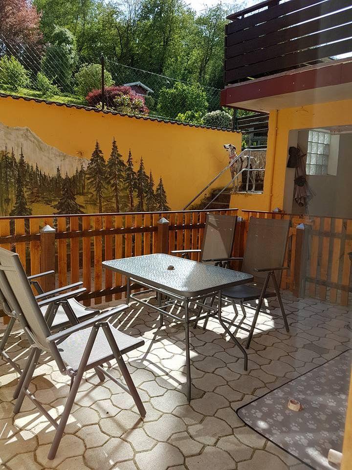 Terrasse mit Tisch und Stühlen, gelber Wand mit Waldmalerei, Holzgeländer und Hinterhof.