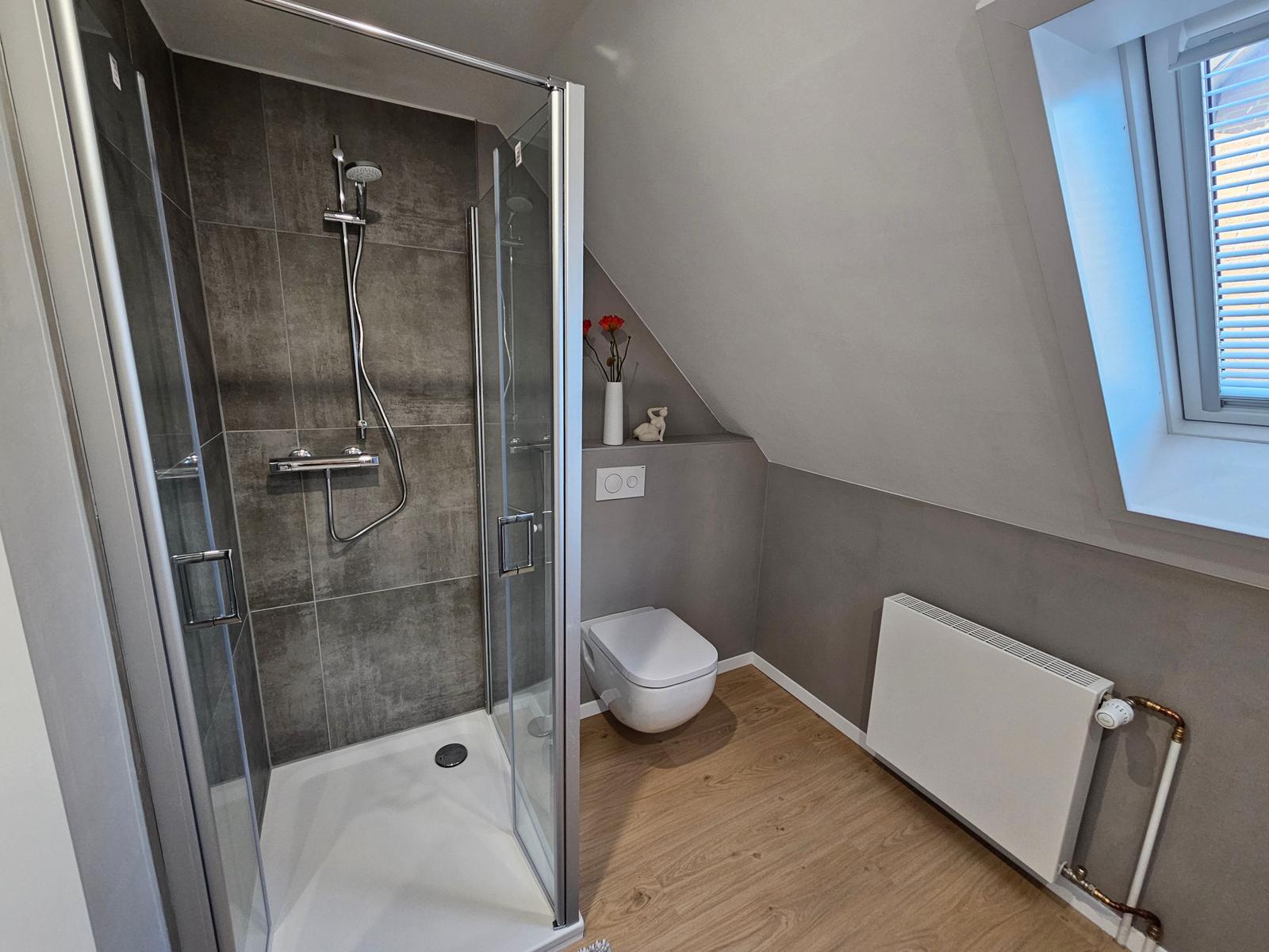 Duschbad mit Dusche, Toilette und Fenster. Holzfußboden und graue Wände.