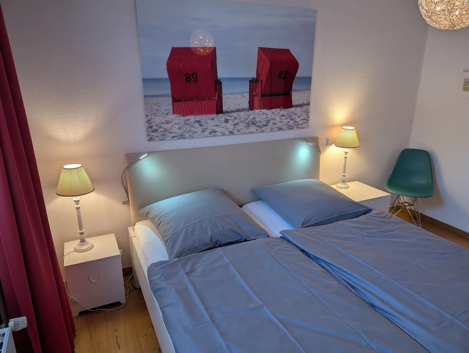 Schlafzimmer mit Bett, zwei Nachttischen, Lampe und Wandbild mit Strandkörben.