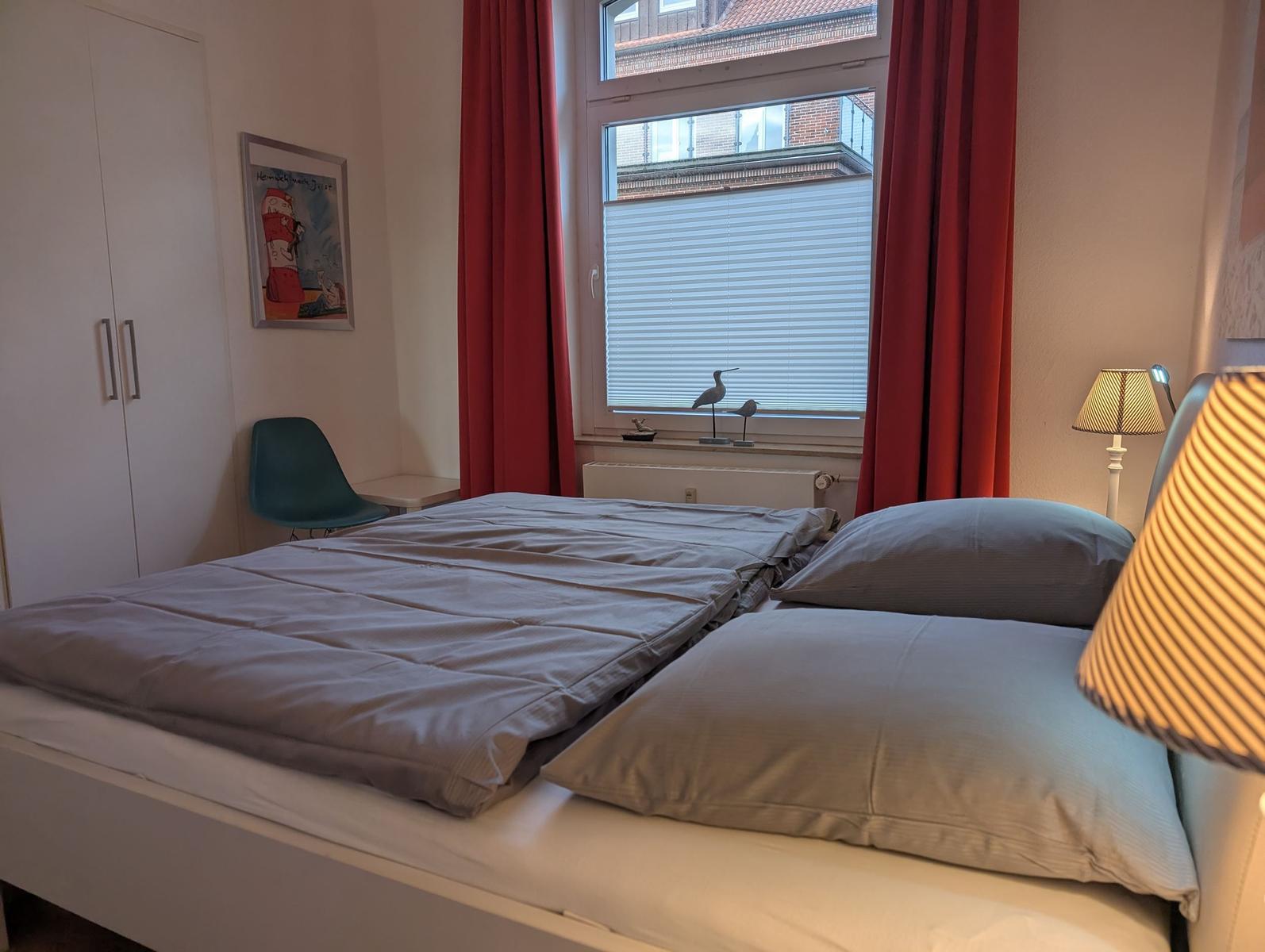 Schlafzimmer mit Bett, roten Vorhängen und Fensterblick auf Gebäude.