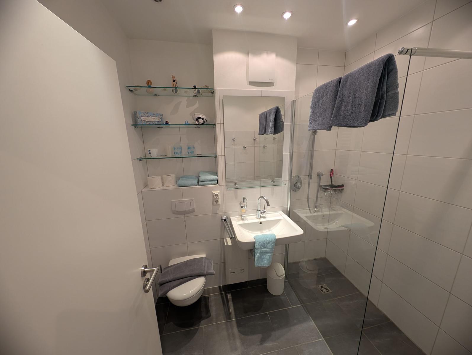 Badezimmer mit Dusche, Waschbecken und Toilette. Weiße Fliesen und graue Bodenfliesen.