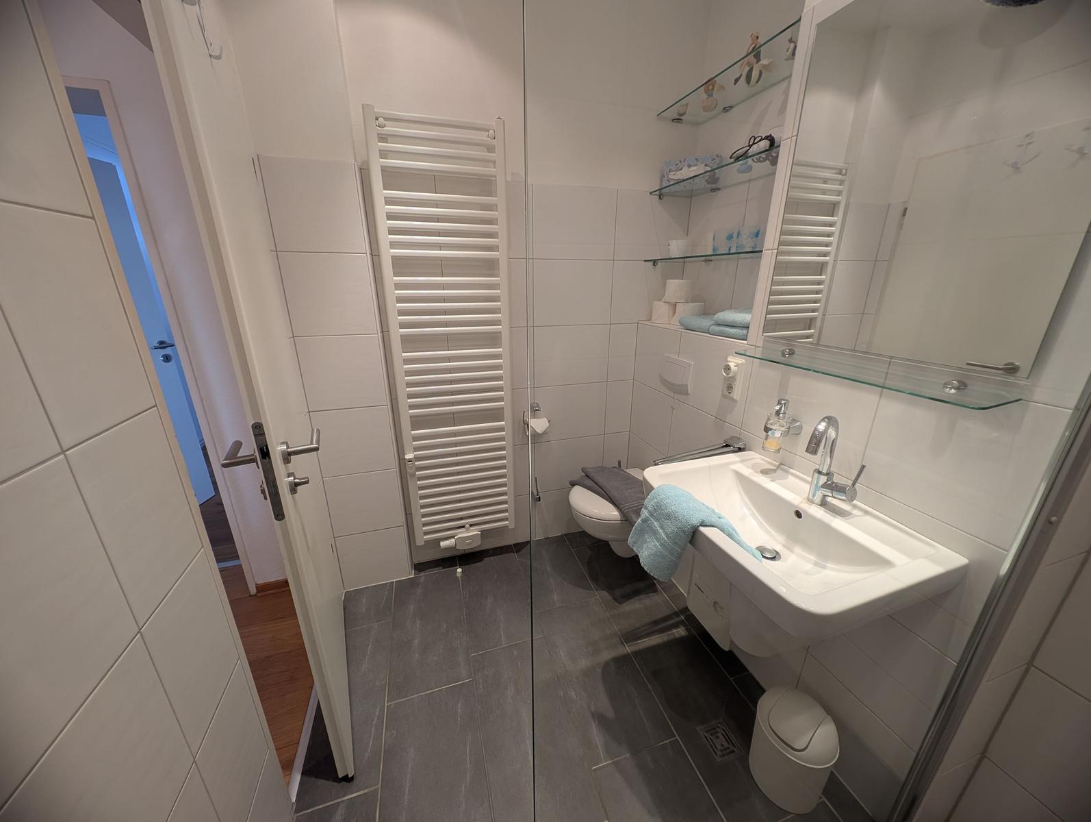 Badezimmer mit Dusche, Waschbecken und Toilette. Weiße Fliesen und graue Bodenfliesen.