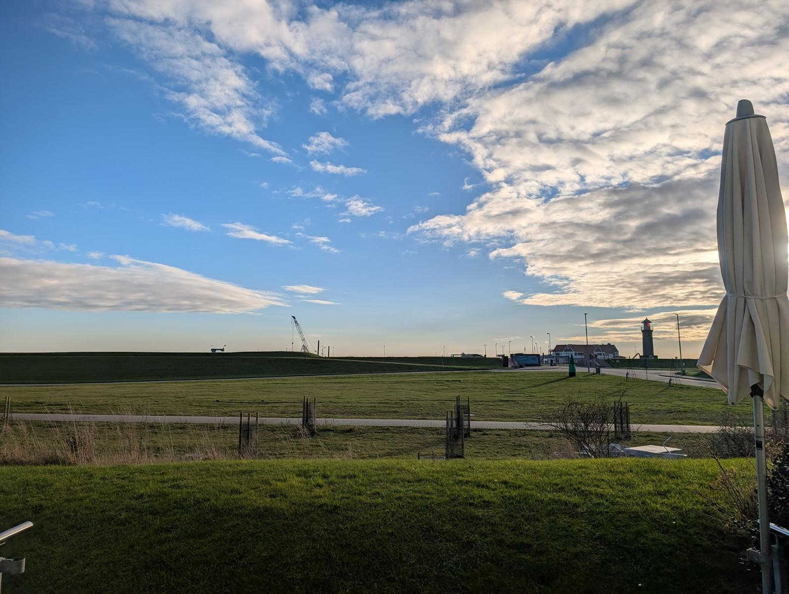 Weitläufiger Grünraum mit Leuchtturm und Sonnenschirm im Vordergrund.