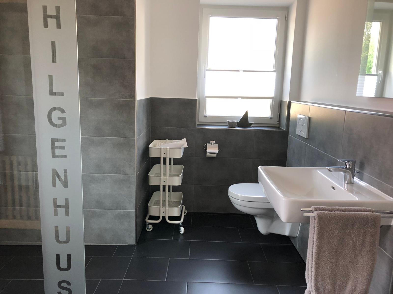 Badezimmer mit Toilette, Waschbecken und Dusche. Graue Fliesen und Fenster.