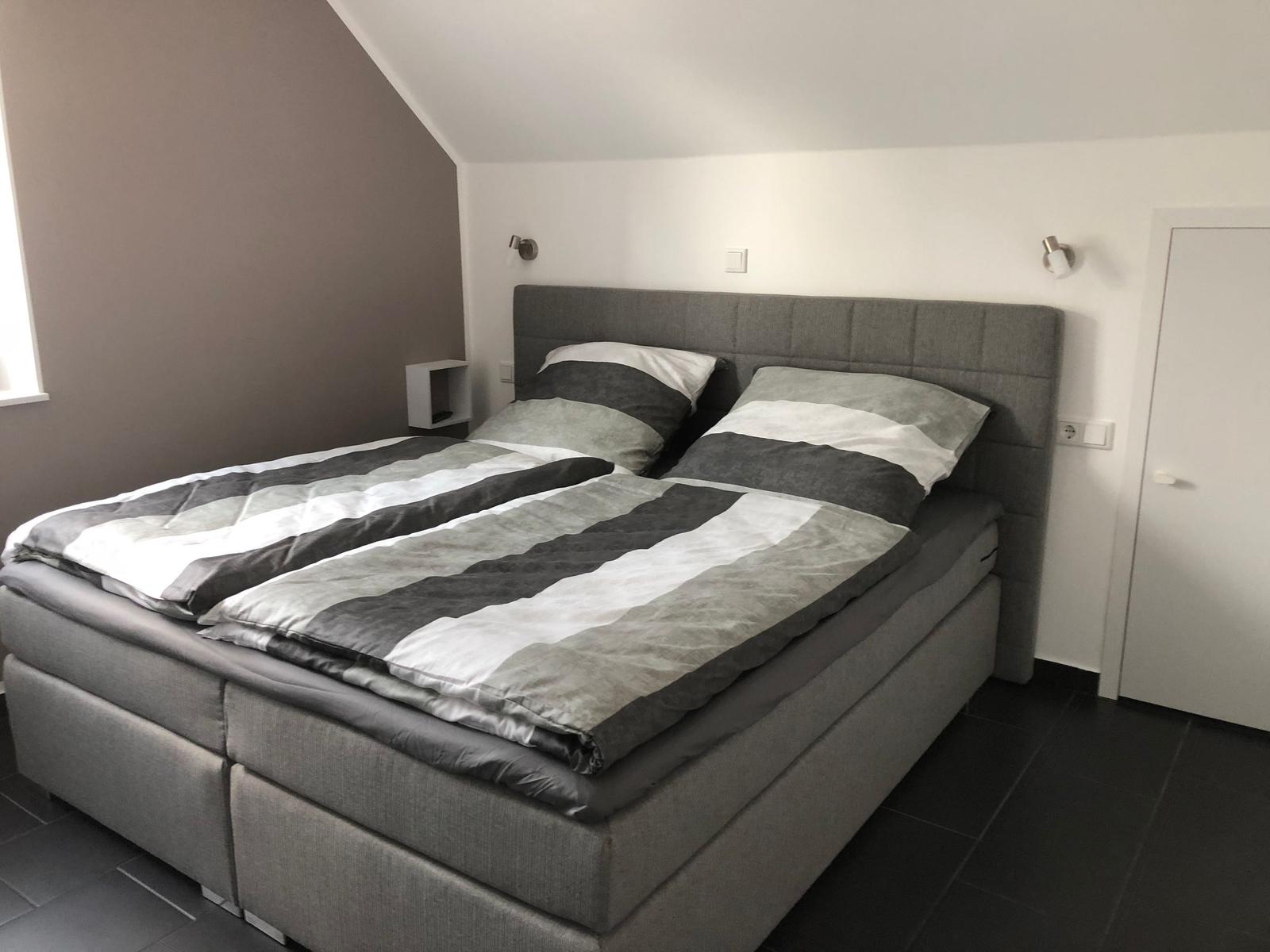 Ein Schlafzimmer mit einem großen Bett und grauer Bettwäsche. Die Wände sind hell und die Decke ist schräg.