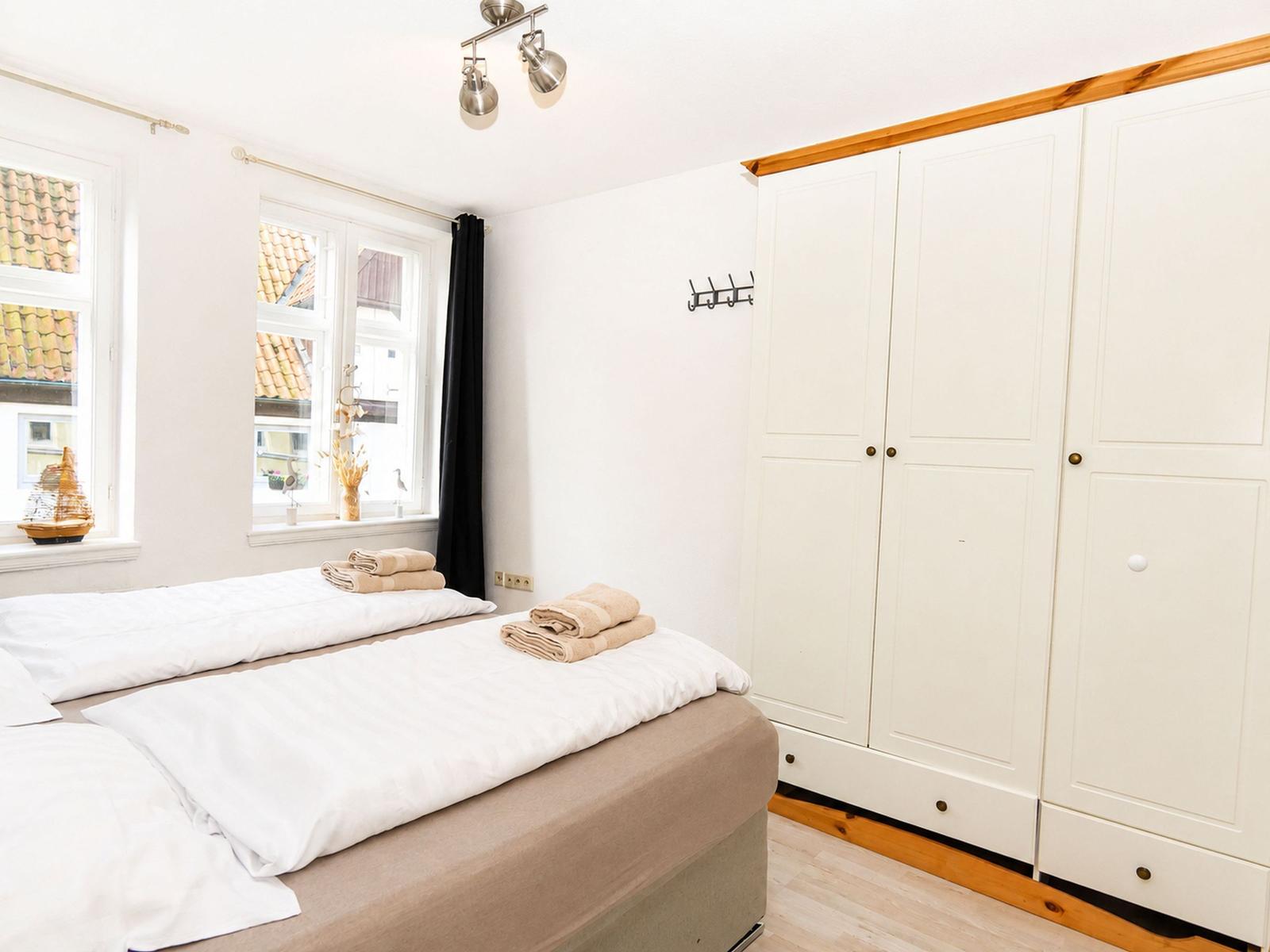 Schlafzimmer mit Doppelbett, weißen Wänden und weißen Schränken. Fenster mit Blick auf Dächer.