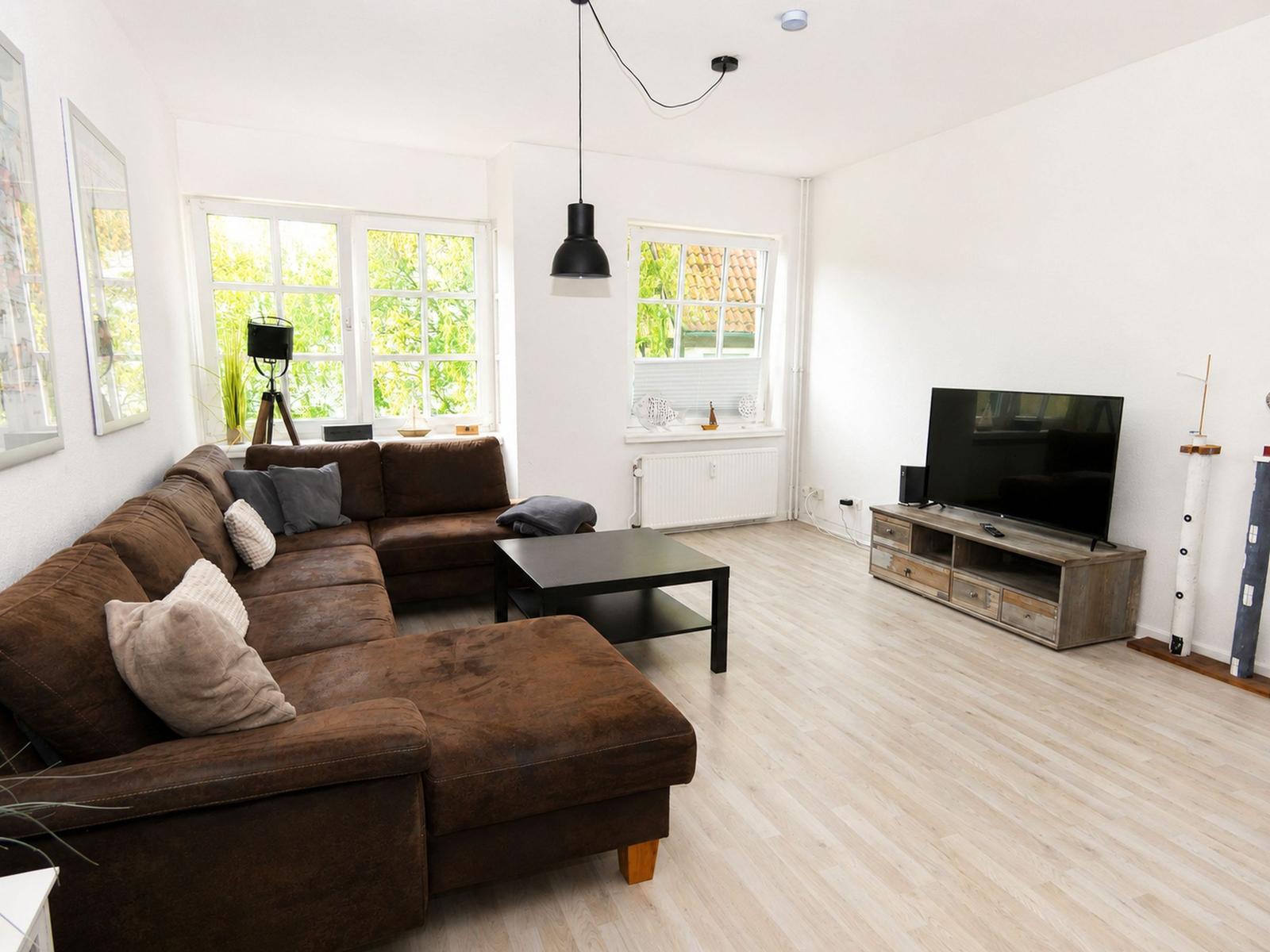 Wohnzimmer mit braunem Sofa, TV und Holzboden. Fenster mit Blick auf Grün.