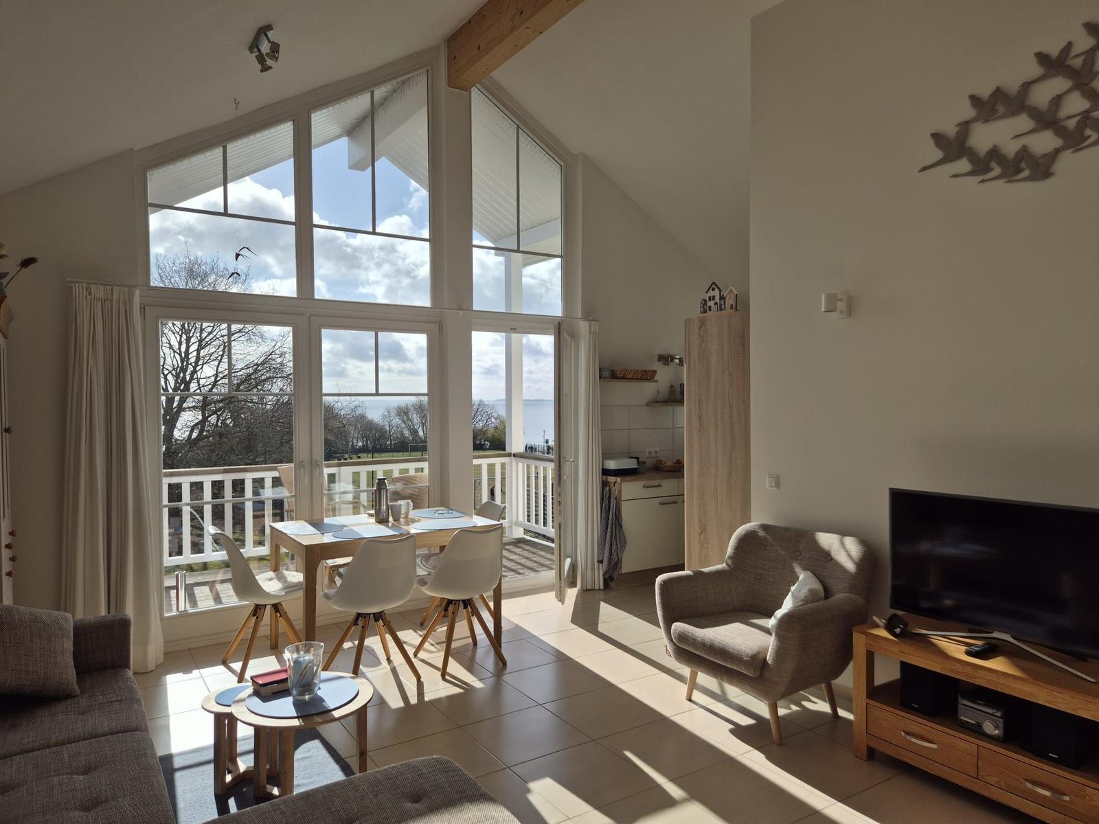 Wohn- und Essbereich mit Blick auf den See und Balkon. Holzdecke, TV und Sessel.