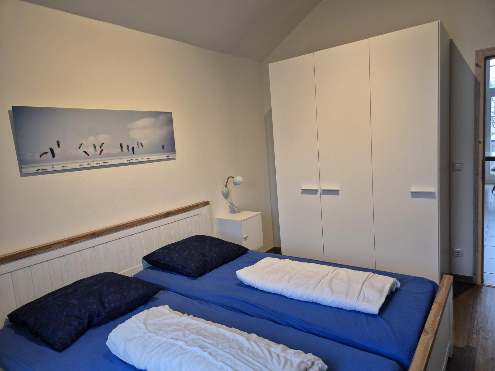 Zimmer mit Doppelbett, Kleiderschrank und Wandbild mit Kitesurfern.