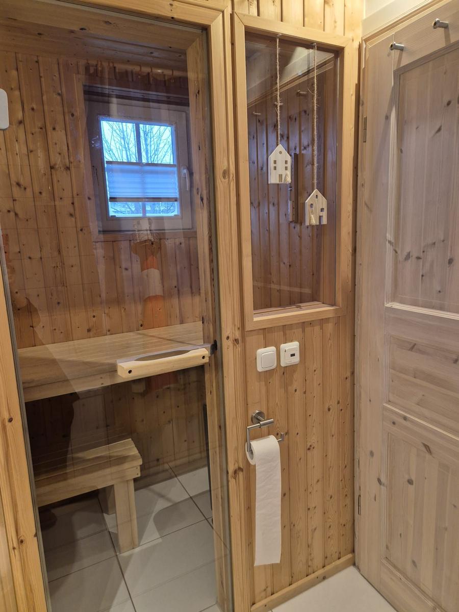 Sauna mit Holzwand, Sitzbank und Fenster. Tür mit Hausanhänger und Toilettenpapierhalter.