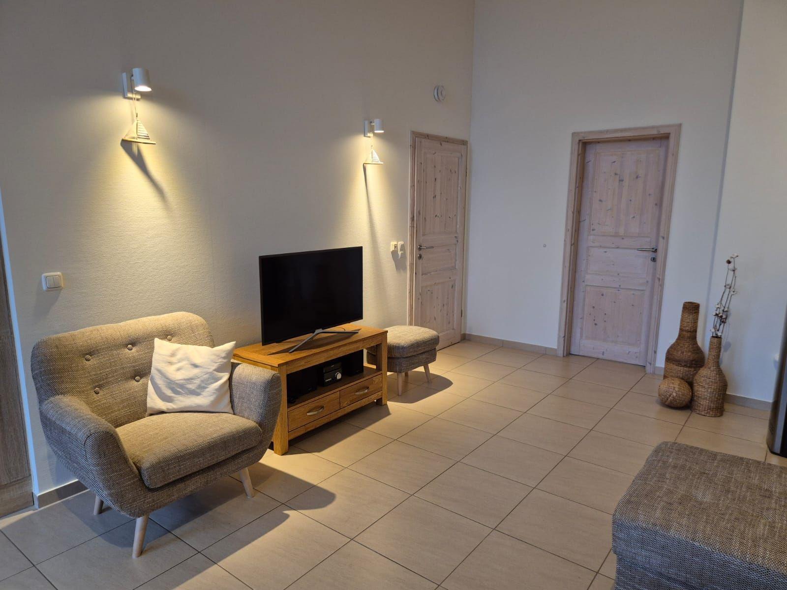 Wohnzimmer mit Sessel, Fernseher und Holztisch. Zwei Türen und dekorative Vasen sind sichtbar.